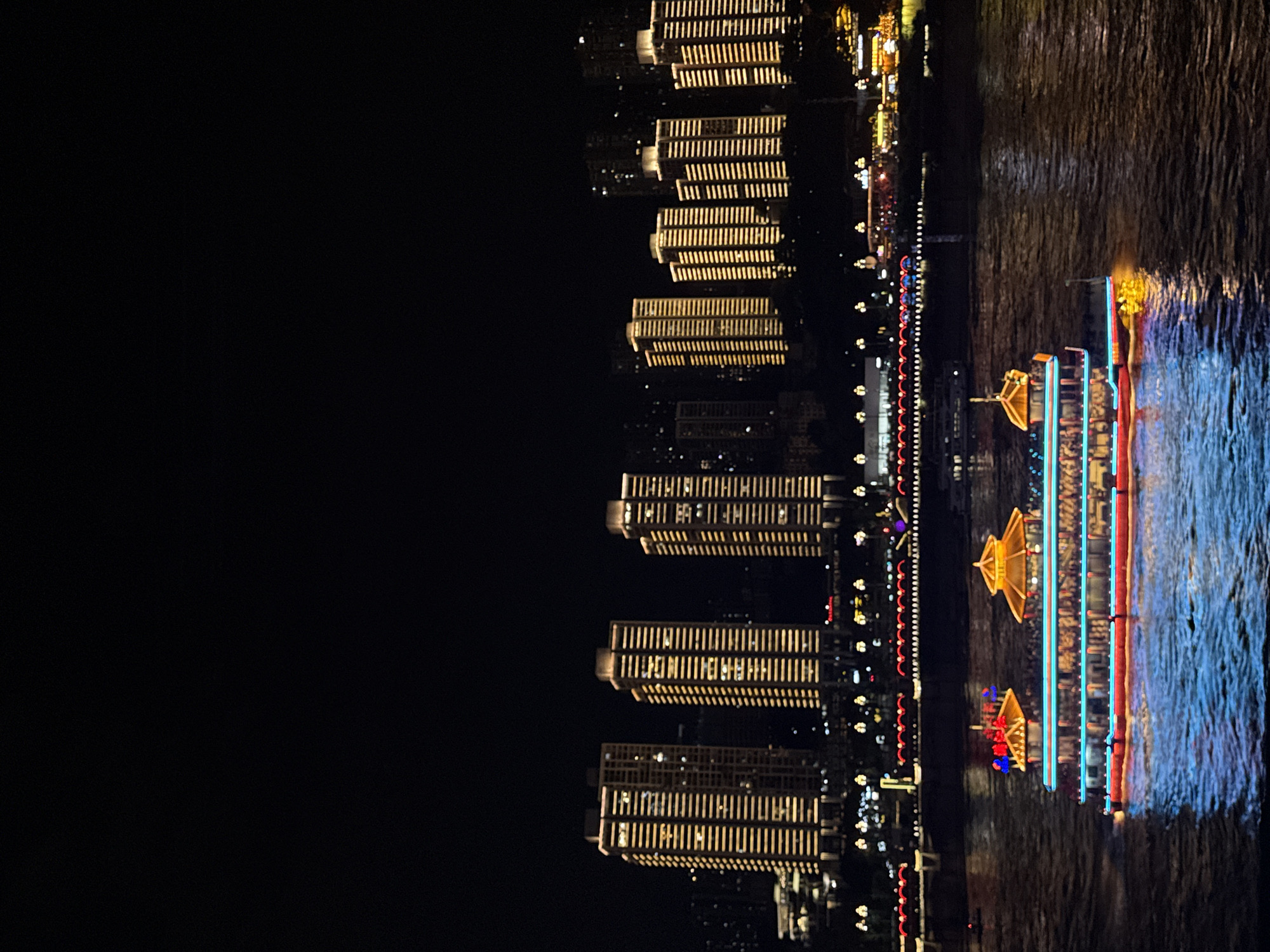 This is a night photograph of the Yuzhong District in Chongqing, China, with a focus on the illuminated cityscape and a brightly lit boat on the Yangtze River. The upper portion of the image is dominated by a dark sky, with the faint outlines of distant buildings visible. Below the sky, a line of tall, modern buildings stands illuminated with rows of warm yellow lights, creating a striking contrast against the darkness. These buildings appear to be office towers or residential complexes.
In the foreground, a multi-decked boat navigates the dark waters of the river. The boat is adorned with vibrant lights in red, blue, and yellow, with a distinct pagoda-like structure on its upper deck. The lights from the boat reflect brightly on the rippling surface of the water, creating a dynamic and colorful display. The boat is carrying passengers, visible as silhouettes behind the illuminated windows. The far bank of the river shows more lights from the city, suggesting a densely populated urban area. The overall scene conveys the lively and illuminated atmosphere of a major Chinese city at night.