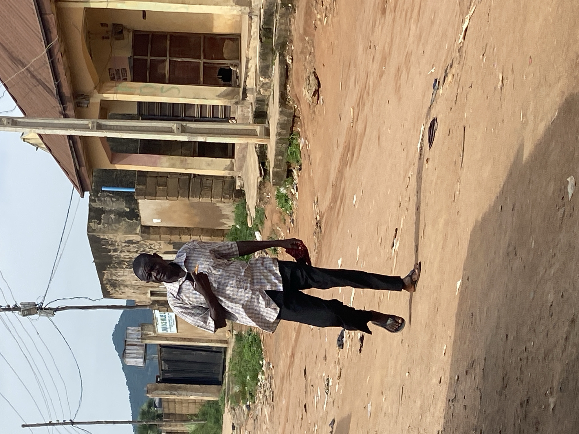 A man walks down a dirt road in Kuson, Nigeria. He is wearing a short-sleeved plaid shirt, black pants, and sandals. He holds a small object in his right hand near his mouth and a red cloth in his left. The road is dusty and littered with debris. In the background, buildings with weathered facades line the street, and overhead power lines crisscross the sky. A distant hillside is visible in the upper left quadrant.
