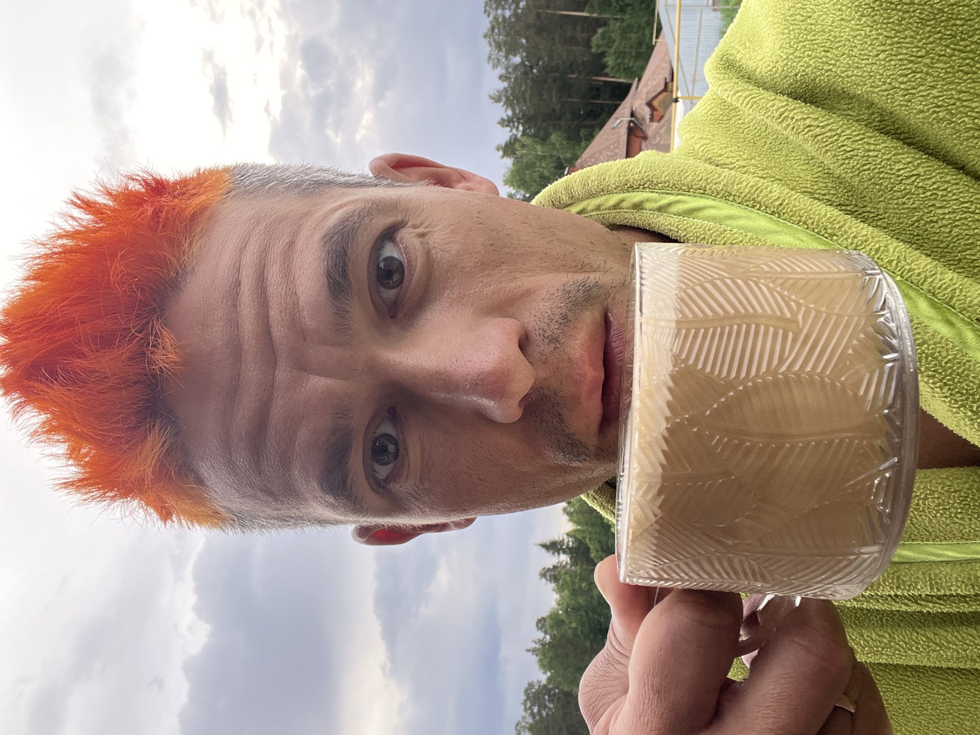 A man with orange hair is holding a mug of liquid up to his face. He is wearing a lime green robe. The background includes cloudy skies, trees, and a building. The location is Chaikovsky, Russia.
