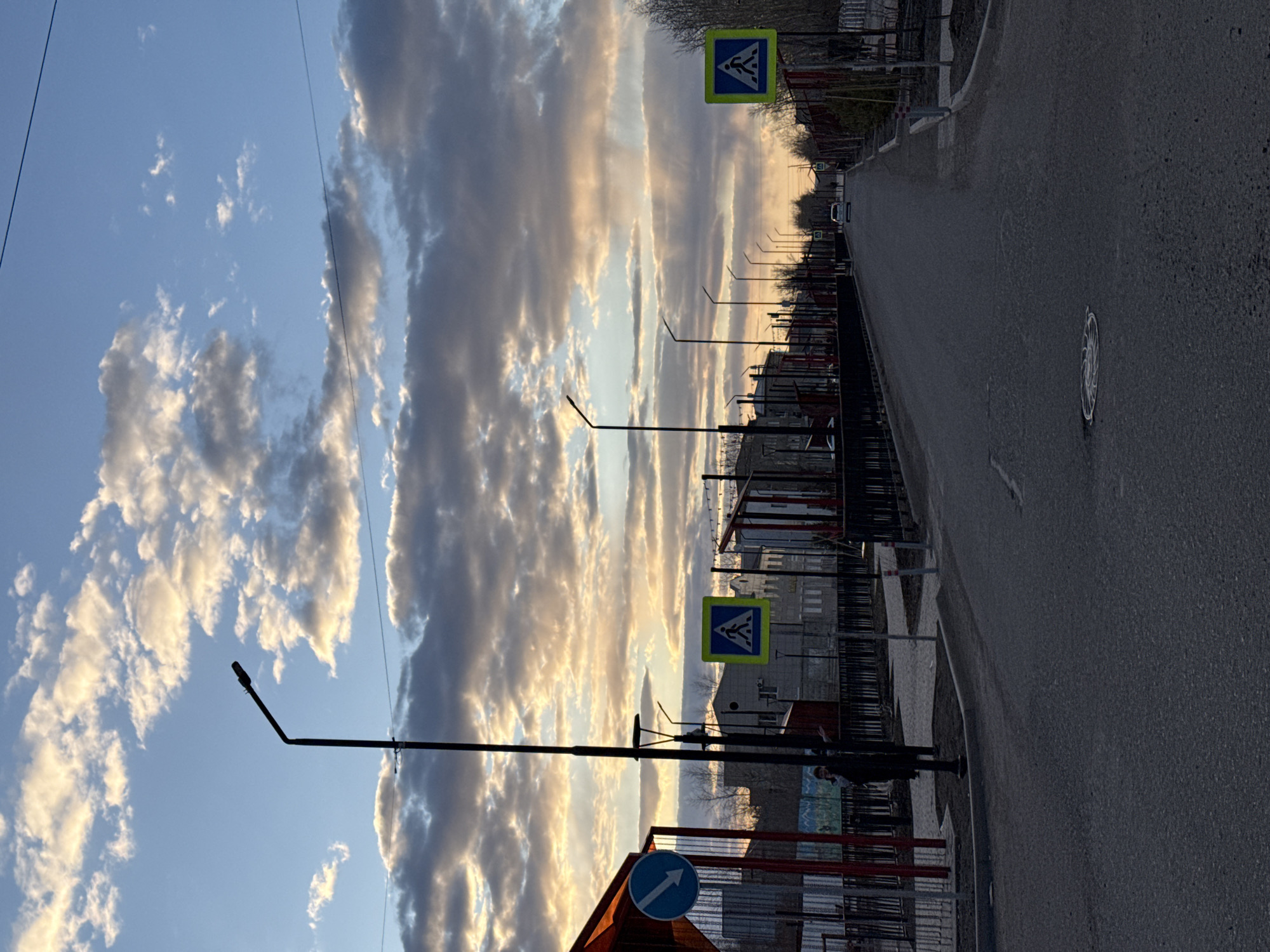 The image displays a street scene in Borzya, Russia, captured during either sunrise or sunset, indicated by the warm, directional light. The photograph is oriented vertically, showing an asphalt road receding into the distance on the right side, flanked by various infrastructure elements.
The sky is a prominent feature, filled with dramatic, elongated clouds that stretch across the frame. These clouds exhibit golden and orange hues where they catch the low sun, contrasting with darker, shadowed sections and patches of clear blue sky. Two thin, dark lines, likely power lines, are visible across the upper portion of the sky.
Along the left side of the road, a line of dark streetlights stands in silhouette against the bright sky, extending into the background. Below the streetlights, dark fences or barriers separate the road from what appear to be buildings or structures and sparse, leafless vegetation. The buildings are largely shadowed, contributing to the sense of early morning or late evening.
Several road signs are visible:
- Two square, yellow signs with a black border and a blue internal panel depicting a white pedestrian crossing symbol, indicating a pedestrian crossing ahead.
- One circular blue sign with a white arrow pointing upwards and to the right, a mandatory direction sign.
The asphalt road surface is dark, and a circular manhole cover with a decorative pattern is visible on the road in the mid-ground. The scene is devoid of visible people or active events, conveying a sense of quietude in an urban or semi-urban setting. The combination of striking cloud formations and the golden hour lighting creates a picturesque yet utilitarian landscape.