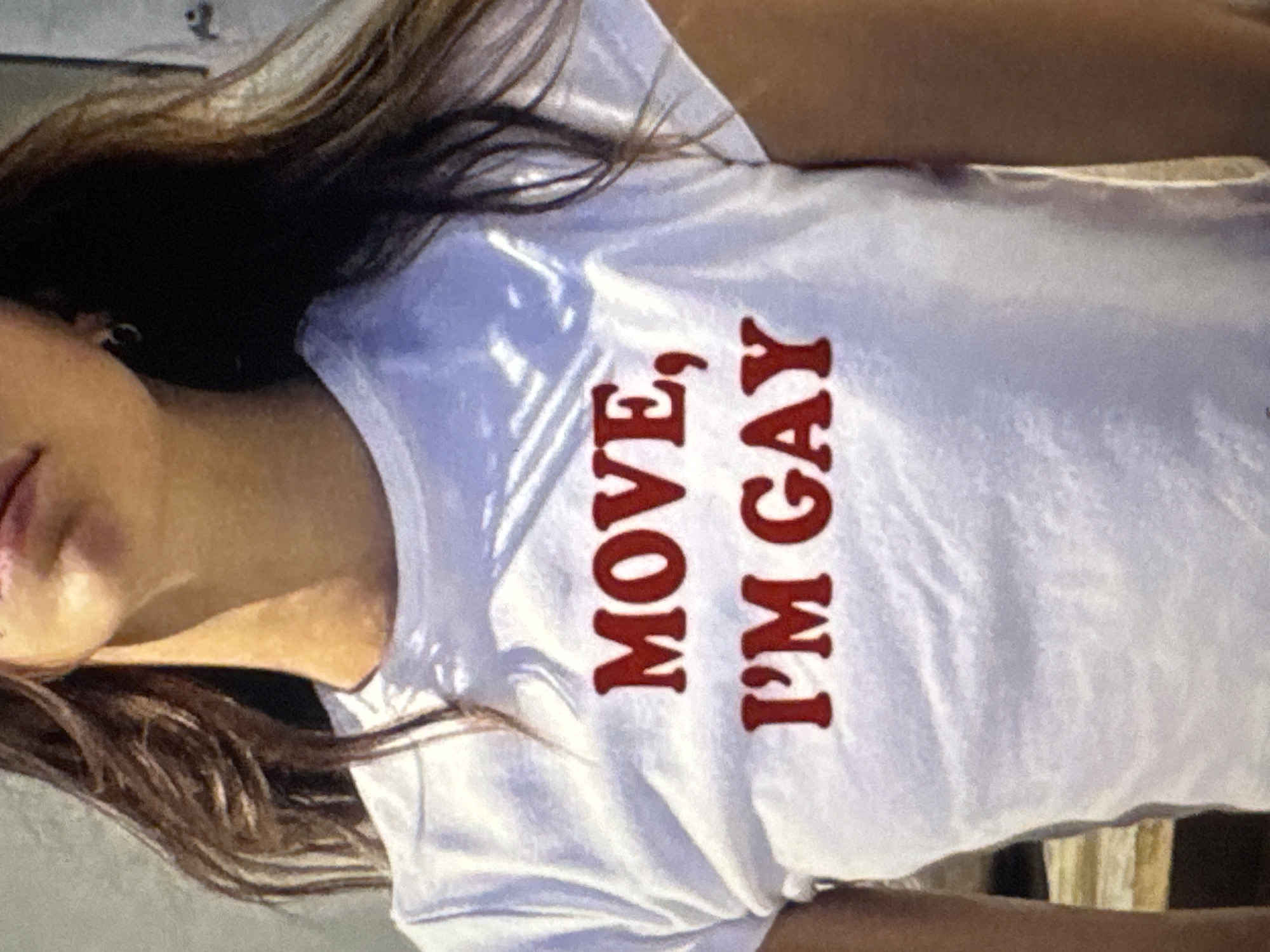 The image shows a close-up of a young person wearing a white t-shirt with red lettering that reads "MOVE, I'M GAY." The person's face is not fully visible, but their hair is dark and appears to be long. The background is out of focus and appears to be a wall with some wooden elements. The location is Kuta Selatan, Indonesia.