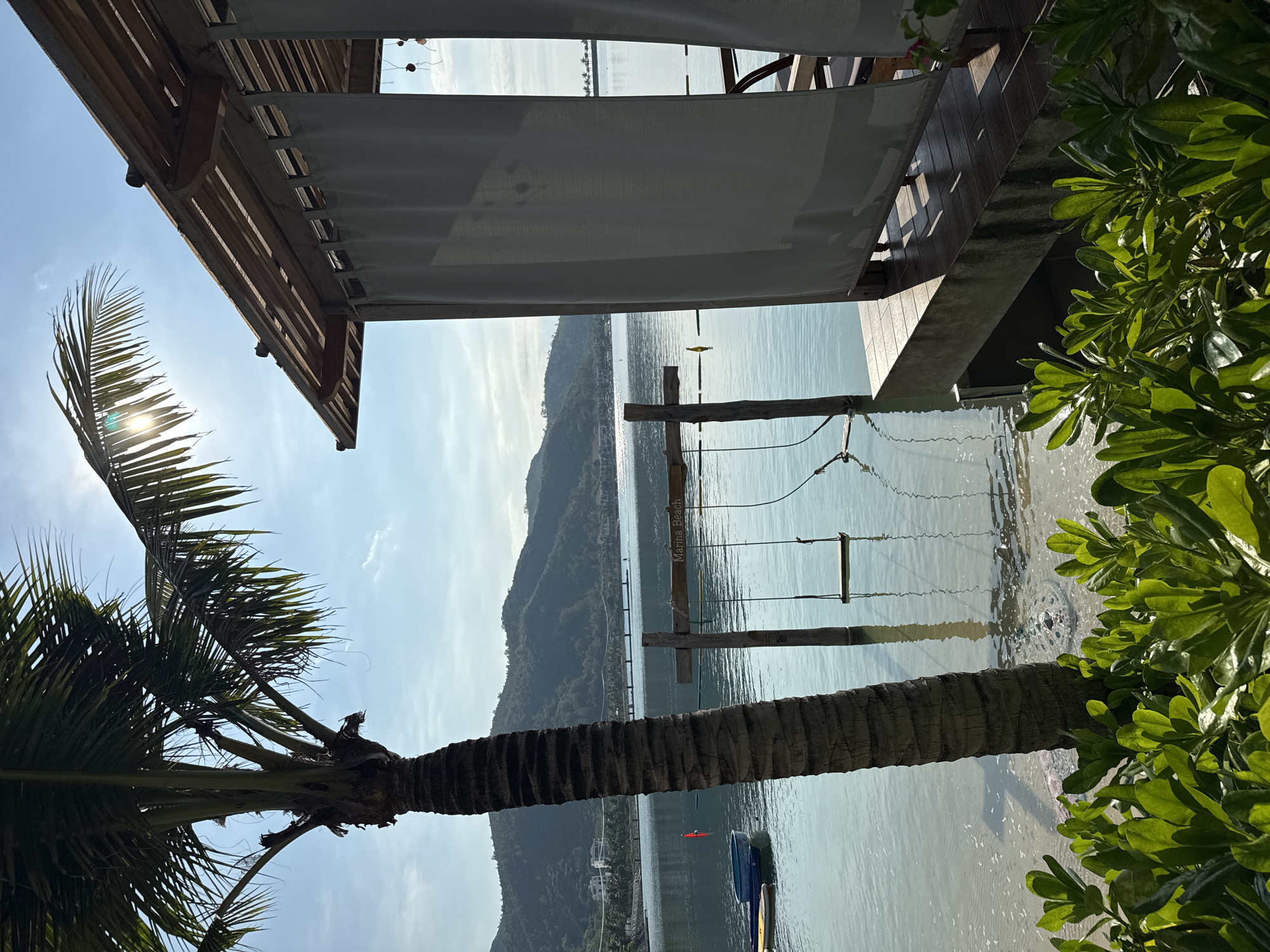 This image depicts a serene waterfront scene in Bac Nha Trang Ward, Vietnam. In the foreground, lush green foliage frames the bottom of the shot. To the left, a tall palm tree stands prominently, its trunk textured and its fronds casting shadows. The calm, shallow water extends from a sandy shore towards the horizon. Partially submerged in the water are two wooden posts supporting a horizontal beam, from which a swing seat hangs. A sign reading "Marina Beach" is attached to the beam. On the right side of the image, a wooden cabana with a slatted roof is visible. Light gray curtains hang from its sides, partially obscuring the interior which appears to have seating. Beyond the cabana and the water, a coastline with rolling green hills and some buildings can be seen under a bright, sunny sky with sparse clouds. The sun's rays are visible, creating a lens flare effect through the palm fronds. A small blue boat and some yellow buoys are also present in the water.