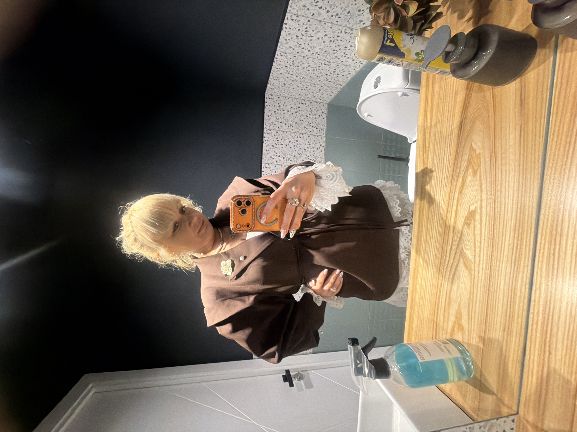 A blonde-haired person is captured in a mirror selfie within a modern bathroom. The individual is shown from the chest up, wearing a stylish brown garment, possibly a coat, with prominent white lace cuffs. They have a neutral expression, adorned with rings on their fingers, a necklace featuring a white flower pendant, and long, decorated nails. An orange smartphone with an embellished smiley-face case is held up to take the photo.
The bathroom setting includes a light wooden countertop where a dark grey pump dispenser, a yellow and white aerosol can, and some artificial plant decor are placed. The aerosol can clearly displays Cyrillic text including "РАБОТАЕТ" (WORKS) and "ЭФФЕКТИВНО" (EFFECTIVELY), along with a floral logo. A clear spray bottle containing blue liquid is also visible on the counter closer to the camera. The reflection in the mirror shows a white toilet and a light blue tiled wall behind it. The wall behind the person (not reflected directly) appears dark, while a speckled grey and white surface (possibly terrazzo-style wall or floor) is also visible. A white door with geometric paneling is reflected in the lower left portion of the image. The scene suggests an indoor setting in Chaikovsky, Russia, with artificial lighting.