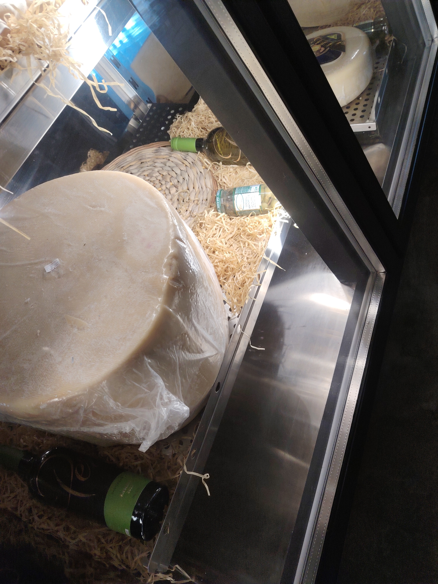 The image shows a display case with various food items and bottles, likely in a store. In the foreground, a large, round cheese wrapped in plastic is prominently displayed on a bed of straw-like material. To the left of the cheese, a dark green bottle with a green label is partially visible, also resting on the straw. In the middle ground, another bottle with a green cap and label, and a clear bottle with a green label are visible. Behind these, a circular cheese with a blue label is on a metal rack. The display case has a reflective surface on its bottom and sides, indicating it's a refrigerated or chilled display. The overall environment appears to be a commercial setting, possibly a delicatessen or market. There is no indication of the city of Konigil, Uzbekistan, in the image.