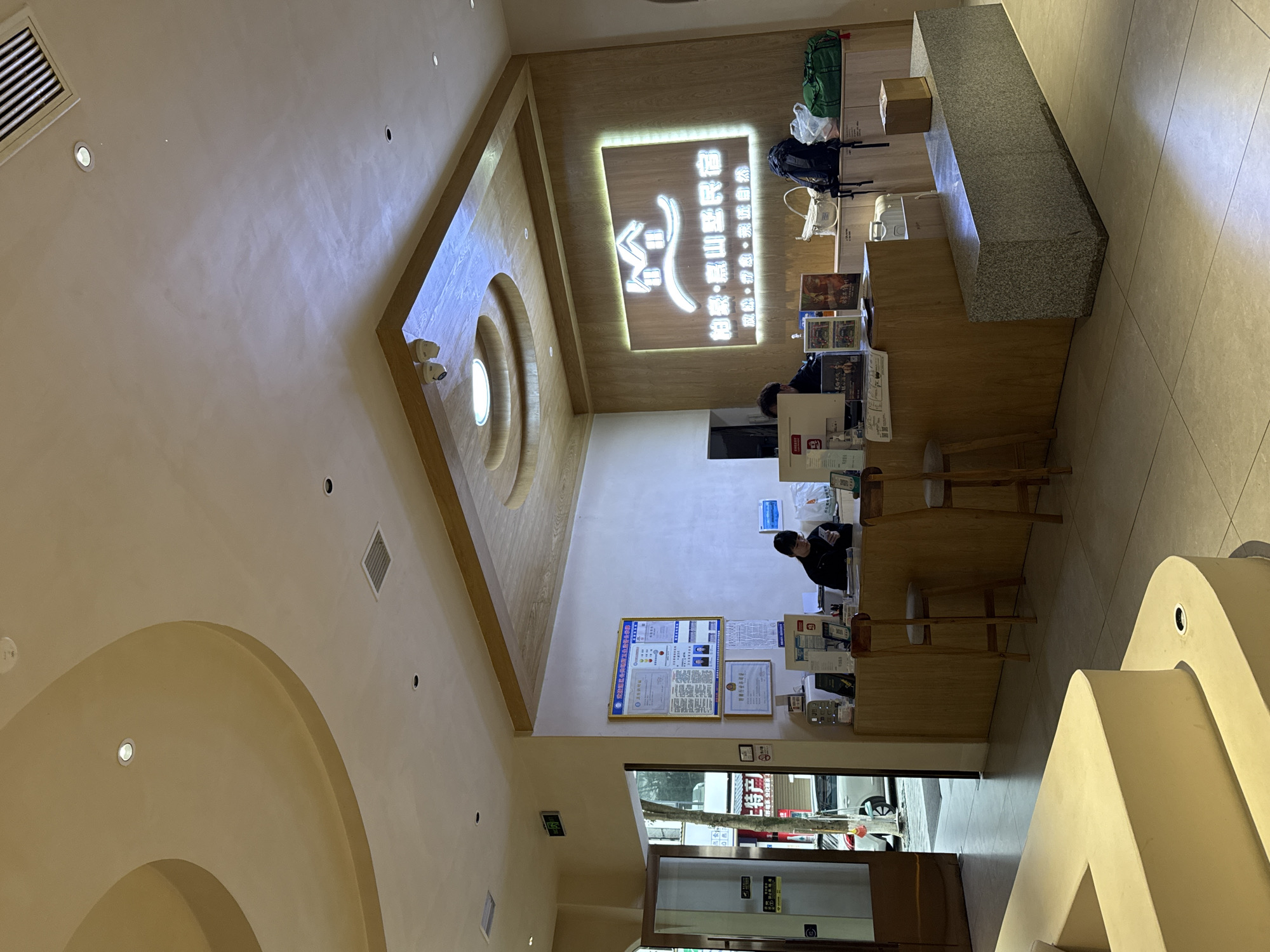 The image displays the interior reception area of the "Baisang Gaoshan Minshu" (柏桑・蒿山亚民宿) guesthouse in Wulingyuan, China. An illuminated sign with the guesthouse name and a two-house logo is prominently displayed on a light wooden wall behind the main reception desk.
Two individuals are present behind a light-colored wooden reception desk. One person, wearing a dark top, is visible on the right, looking down at a mobile device. The other person is partially obscured on the left side of the desk. The desk holds a computer monitor, keyboard, and various documents. Two light-colored wooden high stools with grey padded seats are positioned in front of the desk.
To the left of the illuminated sign, framed certificates and posters are affixed to a light-colored wall. Above the wooden accent wall, a wooden ceiling panel contains a circular light fixture and two white security cameras.
A dark grey granite bench is situated in the foreground, to the right of the reception desk, and supports a brown cardboard box. Various pieces of luggage, including a black backpack, a white tote bag, and a green duffel bag, are placed on a lower cabinet behind the granite bench.
The floor is covered with light beige tiles. The ceiling is light-colored with multiple recessed spotlights, air vents, and a large, curved architectural feature on the left. An open doorway on the left reveals an exterior street scene with a tree, a parked white car, and other buildings. A green exit sign with Chinese characters is mounted above this doorway.