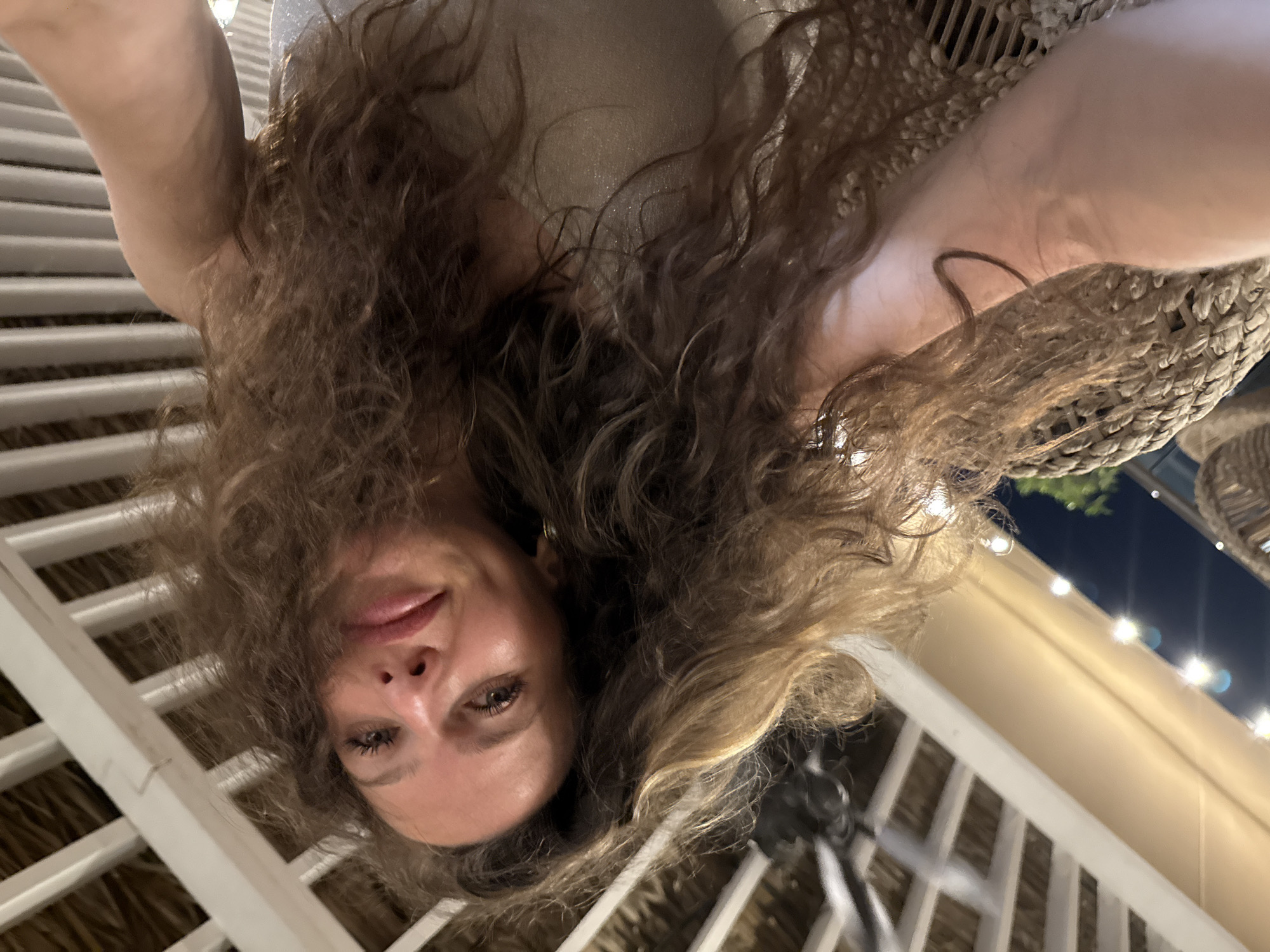 The image shows a close-up, selfie-style shot of a woman with long, curly brown hair. She is looking towards the camera with a slight smile. She is wearing a sparkly, light-colored top. The background appears to be an outdoor setting with a thatched roof structure overhead, supported by white beams. String lights are visible in the upper left corner of the image, suggesting it is evening or nighttime. The woman is seated on a woven chair, with a portion of her arm visible on the left side of the frame. There is no discernible information within the image to indicate a specific location in Dubai, United Arab Emirates.