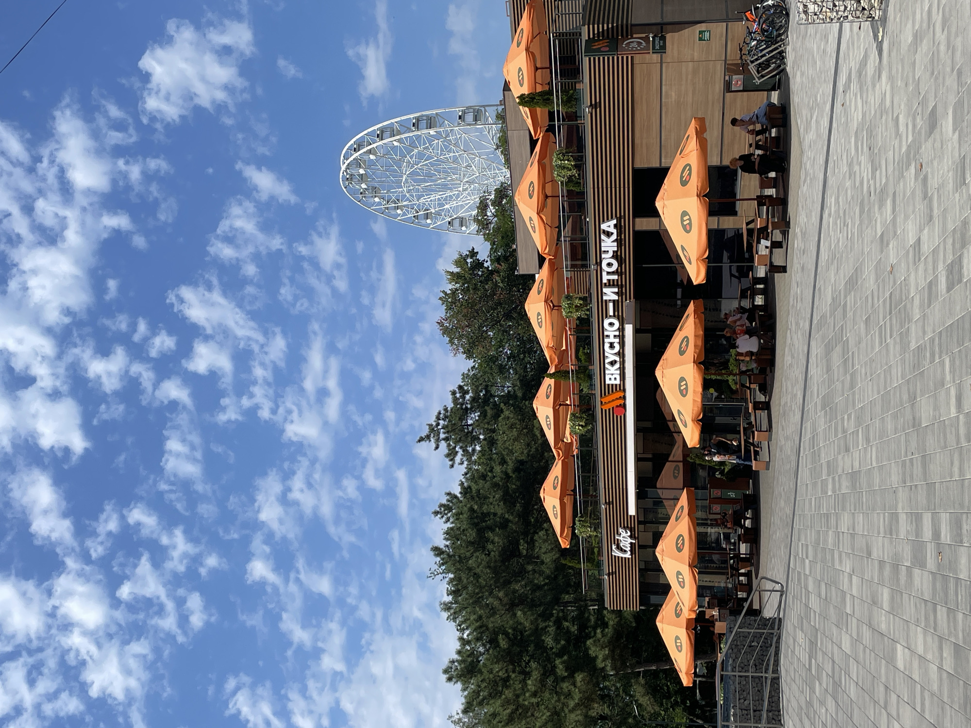In Anapa, Russia, on a sunny day with a sky full of scattered white clouds, a modern cafe with orange umbrellas and outdoor seating is visible. The cafe features a light brown wooden facade with horizontal planks and large windows. The cafe's signage displays "Cafe" in white cursive and "ВКУСНО — И ТОЧКА" in large white letters. In the background, a white Ferris wheel stands prominently against the sky. Lush green trees surround the cafe, and a paved walkway with geometric grey tiles leads to the seating area. Several picnic tables with benches are arranged under the orange umbrellas, with some patrons seated at them. Bicycles are parked to the right of the cafe. The scene suggests a recreational area in the city.