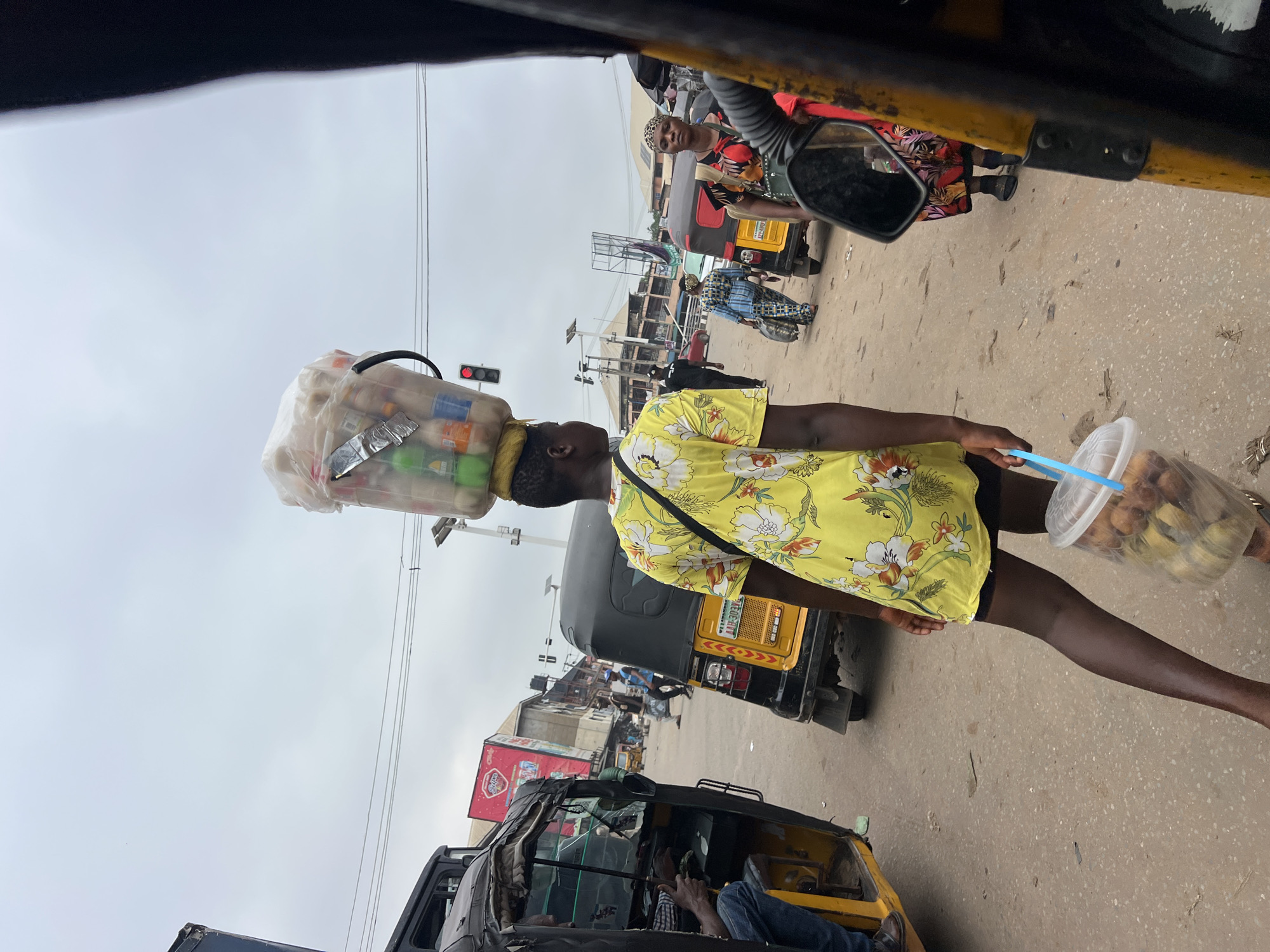 In Nkpor, Nigeria, on a cloudy day, a young woman with dark skin walks away from the camera, her back to the viewer. She is wearing a yellow dress adorned with a floral pattern in white and orange. Her head is topped with a large, clear plastic container filled with various bottles of beverages, possibly juice or water, wrapped in a white plastic bag. She also carries a blue-handled, clear plastic bucket in her right hand, containing several golden-brown fried dough balls, likely puff-puff or doughnuts. Her left leg is bent at the knee, and she appears to be walking on a dirt or asphalt road, which is covered in patches of sand and debris.
The background is a busy street scene. To the left, a yellow and black auto-rickshaw is parked, with a man in a blue shirt and jeans visible inside, seemingly driving. To the right, another yellow and black auto-rickshaw is also present, with a woman wearing a red, black, and yellow patterned dress and a leopard-print headscarf standing beside it, facing forward. Other auto-rickshaws and vehicles are visible further down the street, along with buildings and streetlights. A traffic light, showing a red signal, is mounted on a pole. The overall impression is of a bustling market or transportation hub.