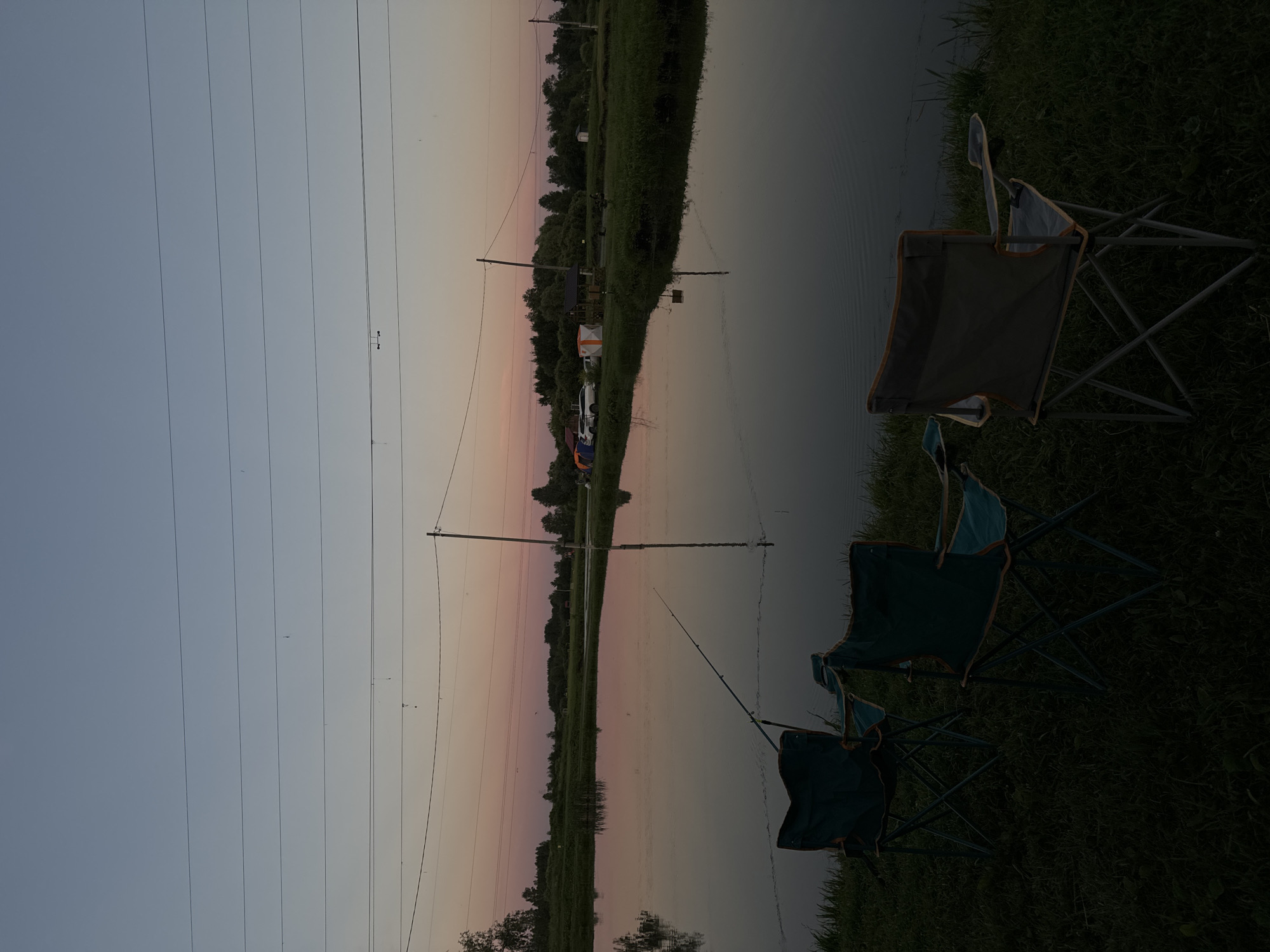 The image depicts a serene waterfront scene at dusk, likely in Komunar, Russia. The foreground is dominated by two dark green camping chairs positioned on grassy terrain. One chair is closer to the camera and slightly angled, while the other is further back and more upright. Beyond the chairs, a calm body of water reflects the dusky sky and the surrounding landscape.
In the middle ground, a thin fishing rod is extended over the water, suggesting an angling activity. Further in the background, a line of trees forms a dark silhouette against the horizon. Among the trees, several tents and a white vehicle are visible, indicating a campsite or a gathering. A tall utility pole with multiple horizontal wires stretches across the frame, dividing the sky.
The sky exhibits a gradient of colors, transitioning from a pale blue at the top to soft pink and orange hues near the horizon, characteristic of sunset or twilight. The overall mood is peaceful and tranquil, with the stillness of the water and the fading light contributing to a sense of calm. No visible text is present in the image.
