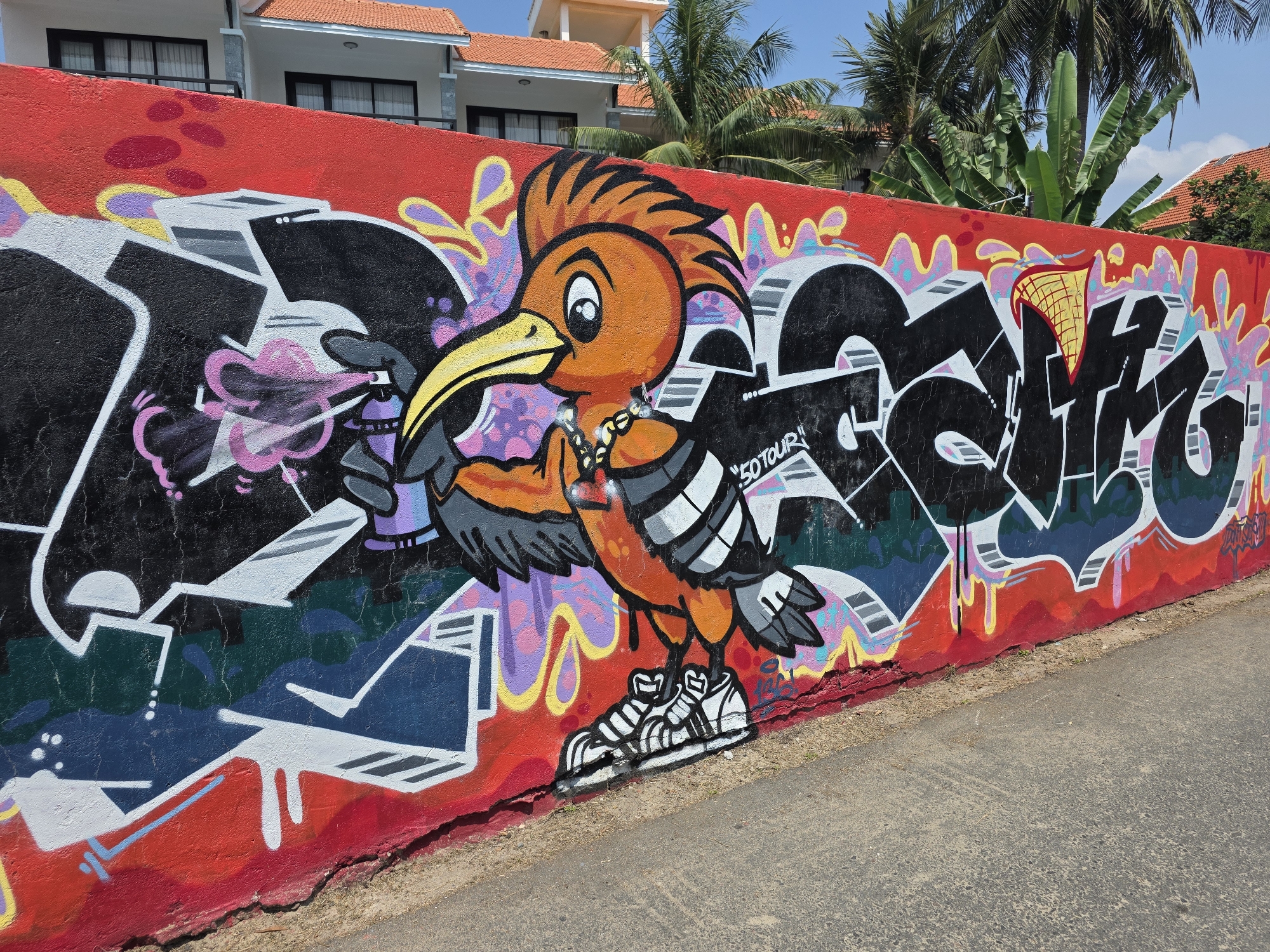 This image shows a colorful graffiti mural on a red wall in Mũi Né, Vietnam. The mural features a cartoon bird wearing sneakers and a chain around its neck, holding a spray paint can. Other abstract graffiti elements surround the bird, including black and white lettering. The background includes buildings with orange tile roofs and palm trees, suggesting a tropical location. A cracked asphalt road runs along the base of the wall.