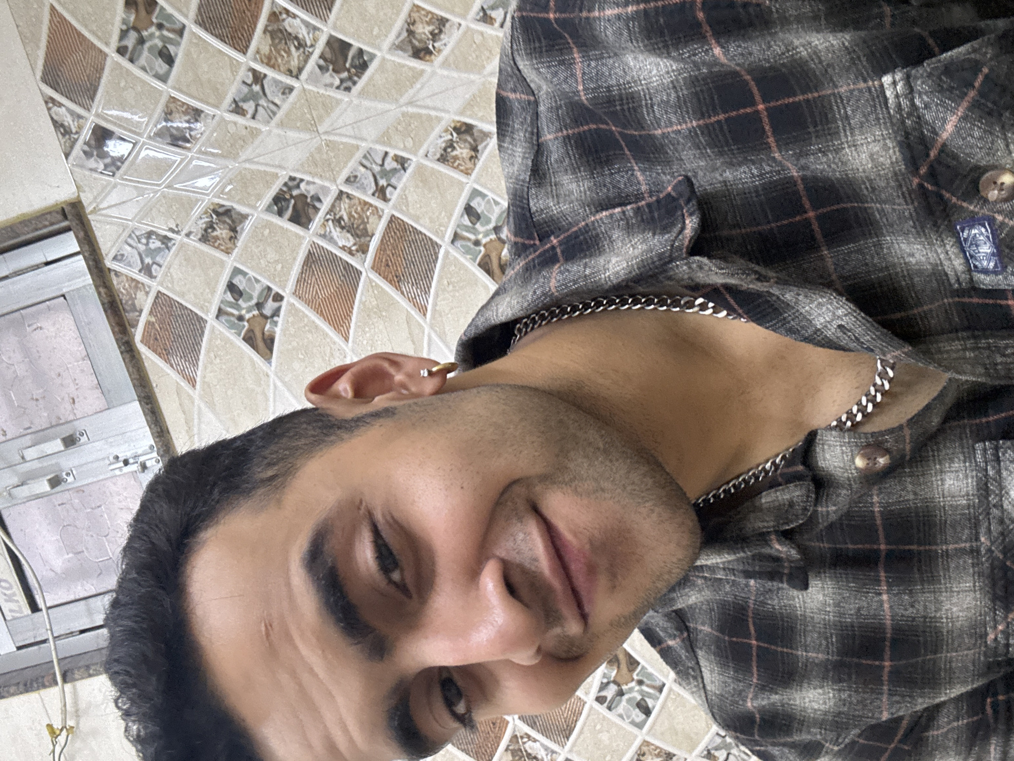 A medium close-up shot captures a man with dark hair and light skin wearing a checkered shirt and a silver chain. The background features decorative tiles, suggesting an interior setting, possibly a bathroom or kitchen. Details like the gold earring and a glimpse of window fittings indicate a personal space. The man is facing towards the camera, smiling slightly. Without additional information, identifying a specific location in Kolkata, India, is not possible.