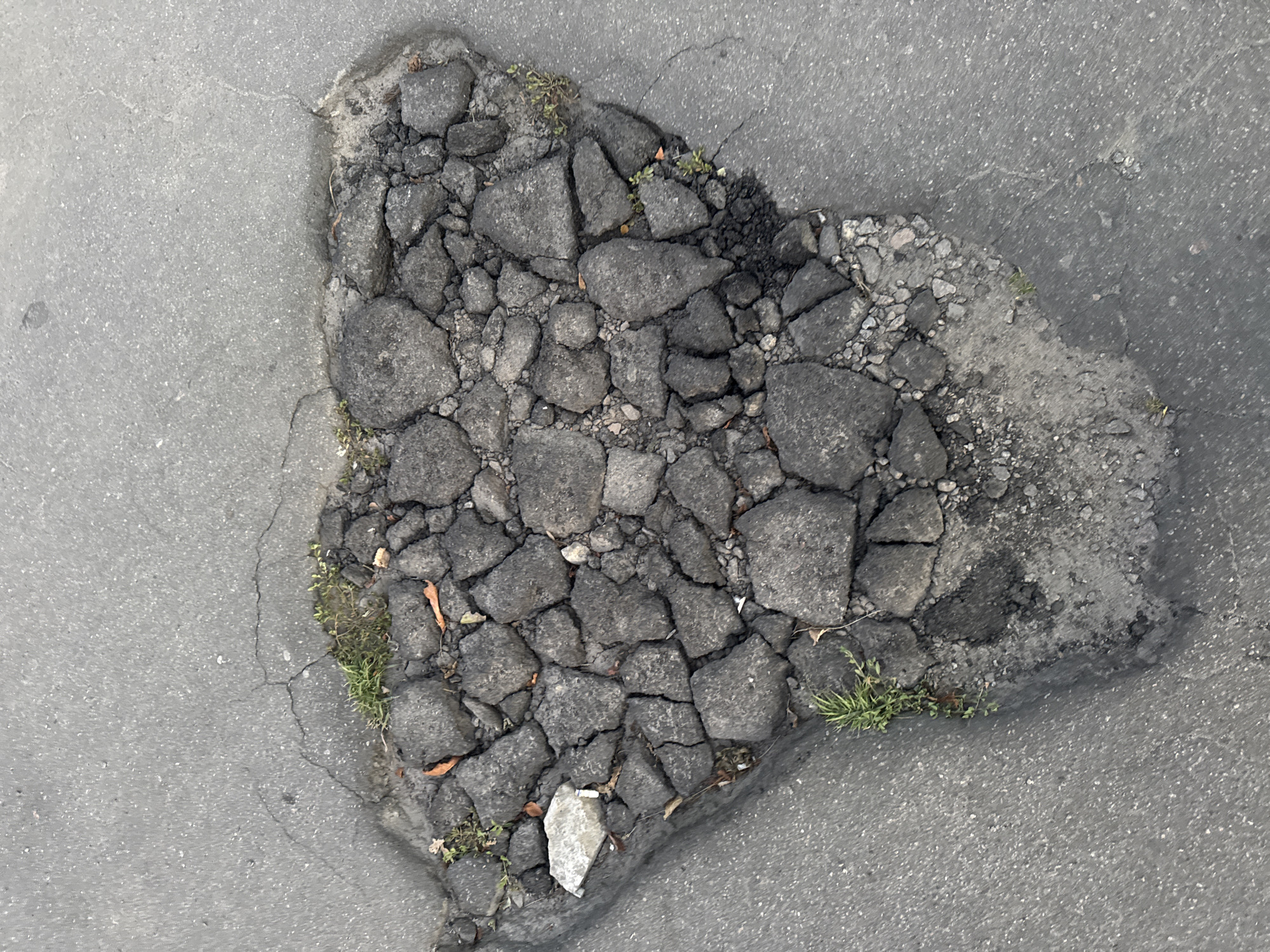 The image shows a pothole in a gray asphalt road. The pothole is filled with broken pieces of asphalt and some small rocks. There are patches of green grass growing in and around the pothole. The asphalt around the pothole is cracked. The scene is consistent with a road in an urban environment. No people or vehicles are visible.