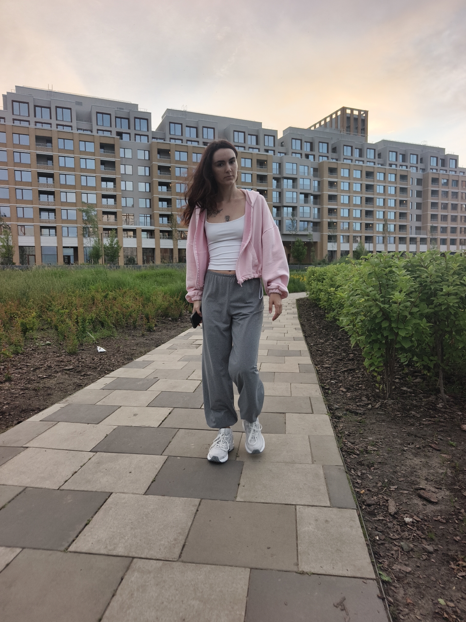 An adult female with dark hair walks towards the camera on a paved path in front of a modern apartment building. She is wearing a pink hooded jacket over a white tank top and grey sweatpants. White sneakers are on her feet. The building is light brown and beige with many windows. To the left of the path is a grassy area with some plants. To the right of the path is a row of green bushes. The sky is overcast with soft, diffused light. The scene is likely set in a contemporary urban residential area.