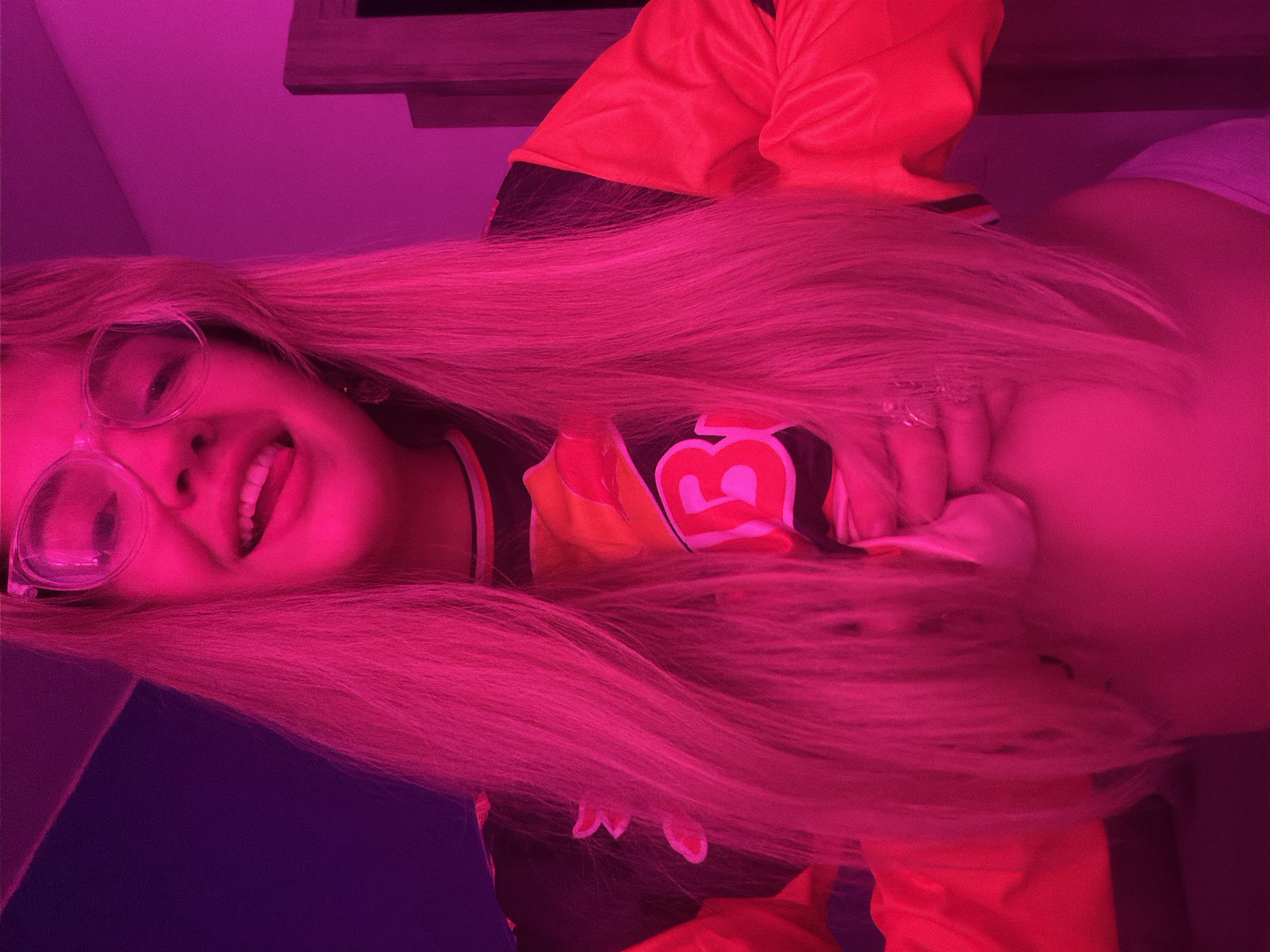 A young woman with long blonde hair and round glasses is smiling and looking at the camera. She is wearing a yellow and black jacket with a large number "3" on the front. The image has a strong pink hue due to the lighting, which makes it difficult to discern specific details of her surroundings. The background is obscured by the pink light, but there appears to be a shelf or cabinet above her and a dark purple wall. The location is Большая Шильна, Russia.