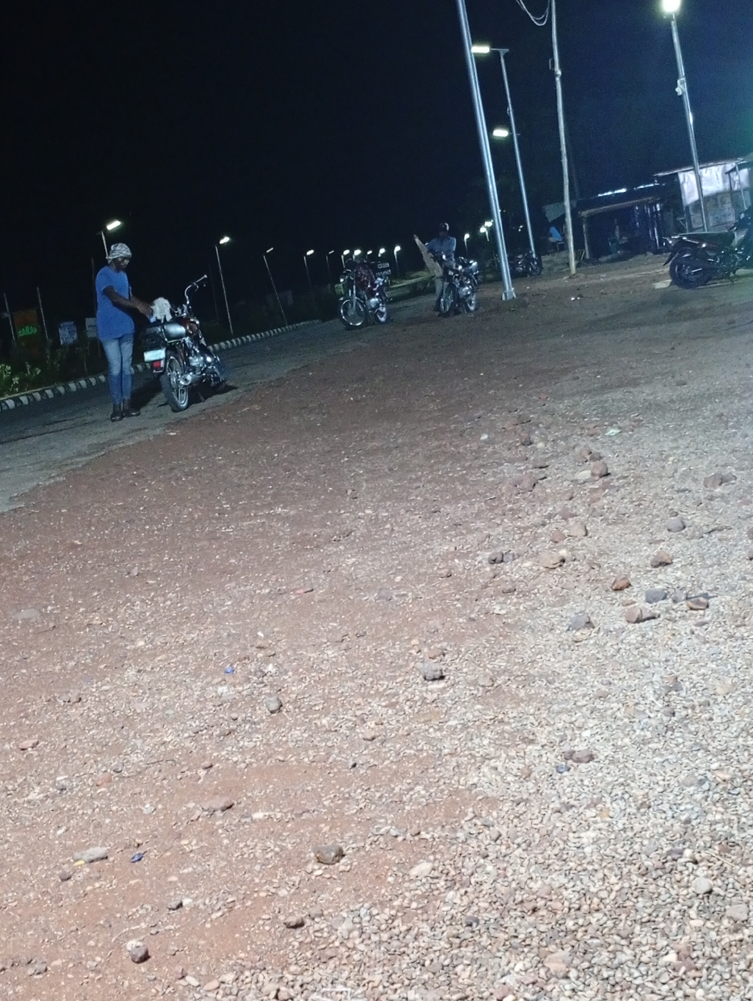 The image depicts a night scene in Adankolo, Nigeria. The ground is an uneven, rocky surface. Several motorcycles are parked under streetlights. A man in a blue shirt and head covering is standing near one of the motorcycles, appearing to be cleaning it with a white cloth. Other motorcycles are parked nearby, and a building with a metal roof is visible in the background.
