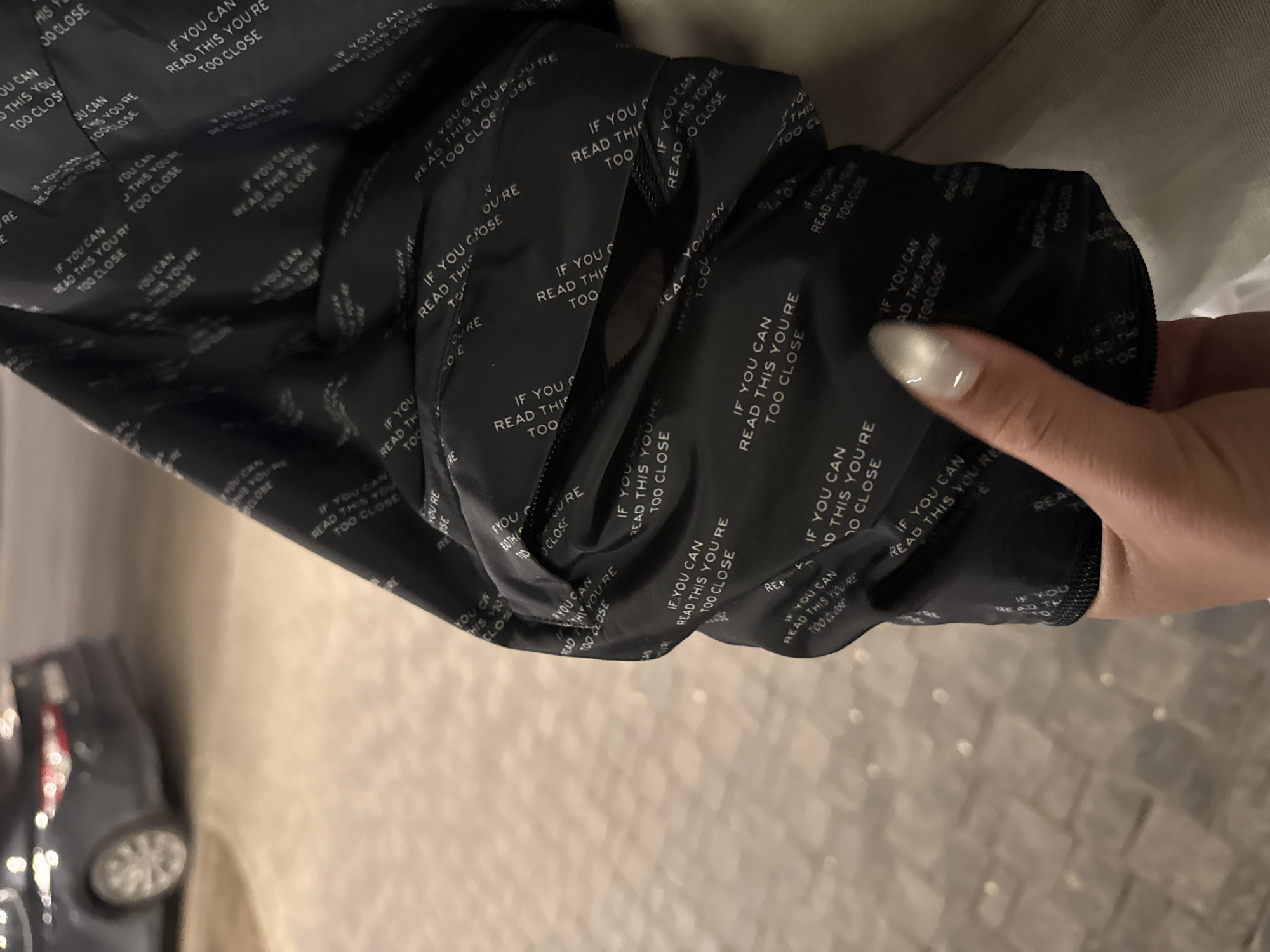 A close-up image shows a person's hand with manicured nails holding and manipulating a piece of black fabric. The fabric, possibly the lining of a jacket or bag, is covered with repeating white text. The dominant phrase visible is "IF YOU CAN READ THIS YOU'RE TOO CLOSE," appearing multiple times, often partially obscured or fragmented due to the fabric's folds and creases. Other visible text fragments include "READ THIS YOU," "TOO CLOSE," "YOU CAN," and "THIS YOU'RE."
The person's fingers, with light-colored, glossy nails, are on the right side of the frame, engaging with the material. The background is softly blurred, depicting an indoor setting with artificial lighting. In the lower-left corner, a dark, blurry object resembling a toy car or model vehicle with visible wheels is discernible on a light-colored floor or surface. The image captures a casual interaction with the textile, highlighting its unique textual pattern. No specific environmental cues confirm the location as Yerevan, Armenia.