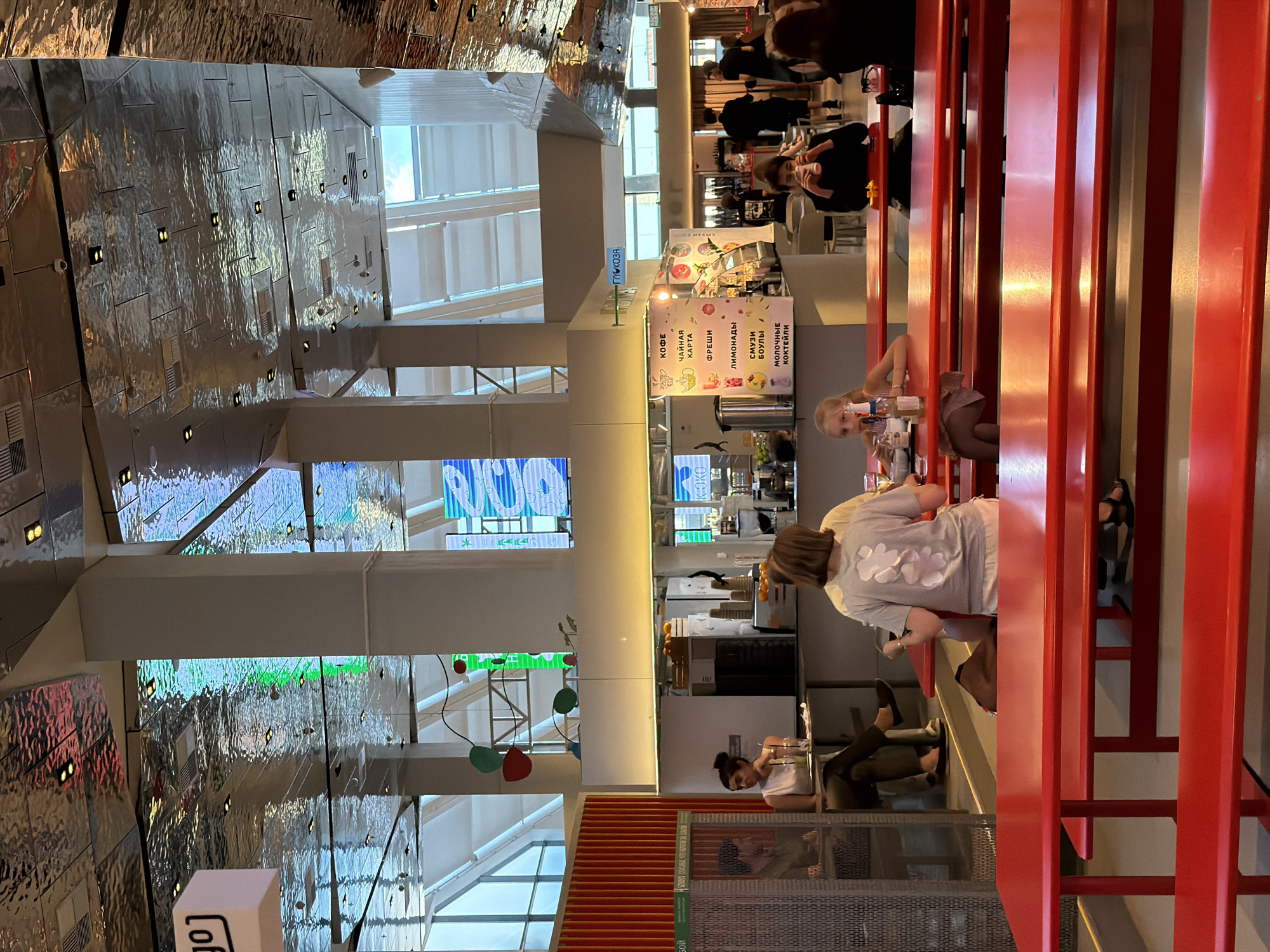 The image captures an interior scene likely within a food court or market in Moscow, Russia. The dominant color scheme is red, seen in the numerous tables and benches that fill the foreground and midground. These tables are arranged in rows, some of which are occupied by patrons.
In the midground, a counter area features a menu board displaying items like "КОФЕ" (coffee), "ЧАЙНАЯ КАРТА" (tea list), "ФРЕШИ" (fresh juices), "ЛИМОНАДЫ" (lemonades), "СМУЗИ БОУЛЫ" (smoothie bowls), and "МОЛОЧНЫЕ КОКТЕЙЛИ" (milkshakes). Behind the counter, there is a large stainless steel beverage dispenser and some crates of oranges, suggesting a juice bar or cafe.
Several individuals are visible. In the left midground, a woman with dark hair tied back is seated at a table, looking towards the viewer, with a drink in front of her. Further back, a person with short blonde hair is seated facing away from the camera, and a young girl with blonde hair is also seated at a red table in the midground, looking to her left. In the background, a few other patrons are seen walking or standing, including a group of people near a food stall.
The ceiling of the space is highly reflective and textured, with a metallic finish that mirrors the lighting and structure above. Large windows or skylights are visible in the background, allowing natural light to enter. The overall impression is a vibrant and modern public space, possibly a market hall or a large shopping center's food court, given the variety of food offerings and the communal seating arrangement. The sign "ГЛЮКОЗА" (Glucose) is visible on a light blue sign hanging from the ceiling.
