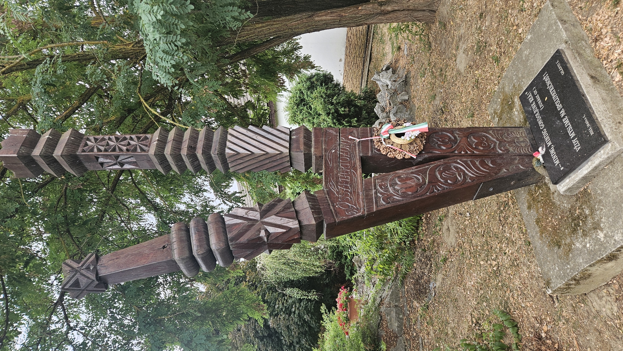 In the city of Veľké Slemence, Slovakia, a wooden monument stands on a concrete base, surrounded by dry grass and trees. The monument is carved with intricate patterns and features a wreath with a Hungarian flag attached. The inscription on the monument's base is in Hungarian, reading: "...MINDIG, MINDIG GONDOLJUNK RÁ!" (JURÁSZ GY.) VZDY MYSLÍME NA SPOLUPATRIČNOSŤ! (GILÁNYI I. 2011, János F.). This translates to "...Let's always, always think of him!" and "We always think of our fellow countrymen!". The overall setting suggests a memorial or historical marker.