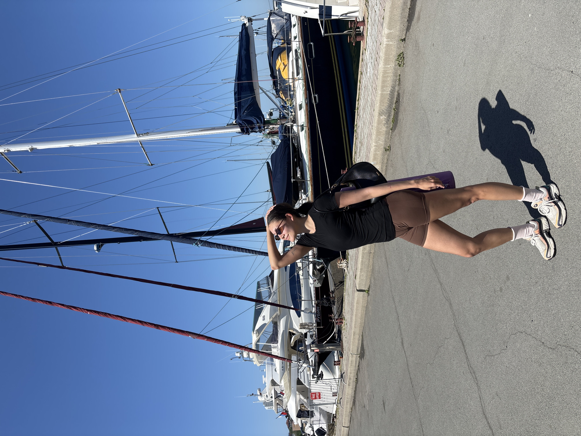 A woman stands on a paved area adjacent to docked boats, likely a marina, on a sunny day. She wears sunglasses, a black shirt, and brown shorts, and carries a yoga mat. Her shadow is visible on the pavement. The background features various boats, including a dark blue sailboat and white yachts, against a clear blue sky. The location is Vladivostok, Russia, indicated by a sign in Russian visible on one of the boats.