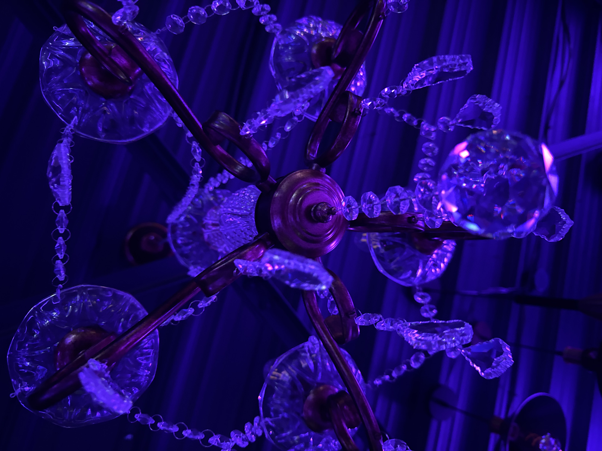The image shows a chandelier with crystal ornaments and a metal frame. The scene is dimly lit, with a predominantly blue hue. The background appears to be draped with fabric. The image shows an upward view of the chandelier. There are no people visible. The location in the city of Créteil, France is not discernible from the visual information provided.