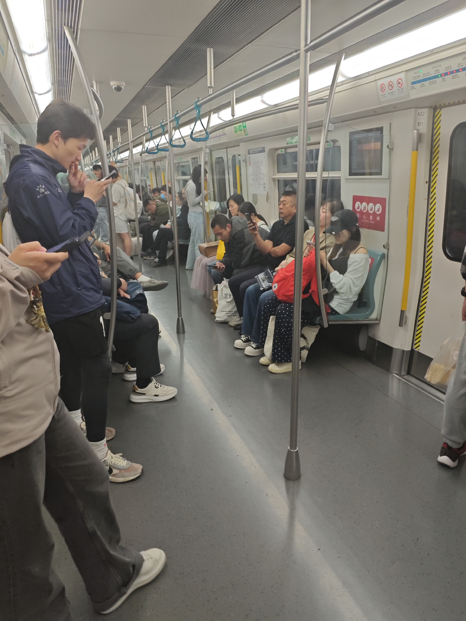 The image shows the interior of a subway car. Passengers are seated and standing. Several passengers are looking at their phones. The interior of the car has silver handrails and overhead lights. Advertisements and subway maps are visible on the walls of the car. The location in the city of Unknown, Unknown, is unknown.