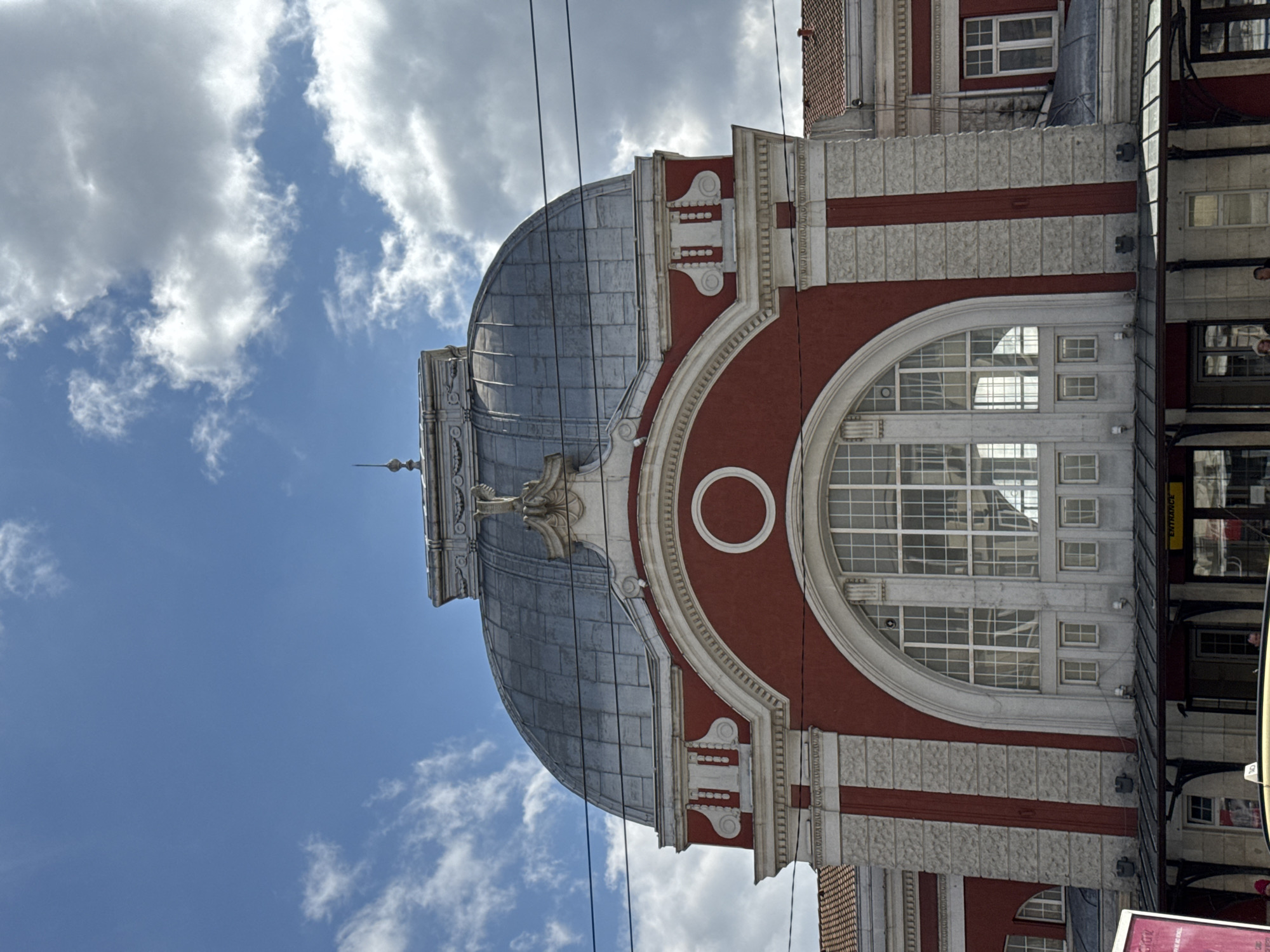 The image shows the Varna Central Railway Station in Varna, Bulgaria. The architecture is characterized by its red and white color scheme, a prominent dome, and large arched windows. The sky behind the station features a mix of blue and scattered clouds. Power lines are visible in the foreground, running diagonally across the frame. A portion of a commercial area is visible on the right side of the image, marked by storefronts, lighting fixtures, and signage including an “entrance” sign.