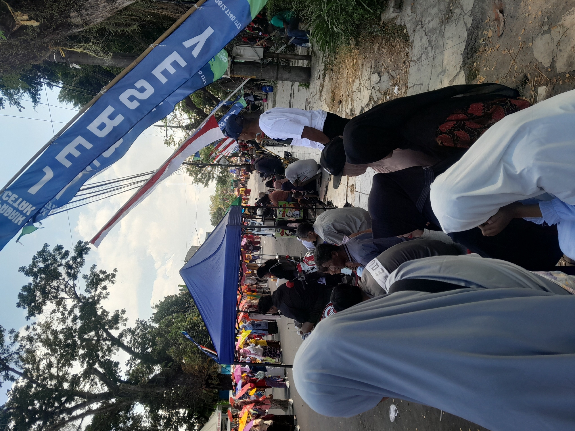 The image depicts an outdoor scene in Genteng Kulon, Indonesia, likely during a public event or gathering. In the foreground, several individuals are seated or standing, facing away from the camera. Many are wearing hijabs, indicating a cultural context. To the left, a blue canopy tent is set up, suggesting a stall or temporary structure. Beyond the tent, a crowd of people can be seen holding colorful umbrellas, indicating fair weather or a festive atmosphere. In the background, several large blue and white banners with text are displayed, likely promotional or political. Trees line the street, and utility poles are visible. The ground is paved and appears to be a street or walkway. The overall impression is one of a busy, public gathering in a tropical or subtropical setting.