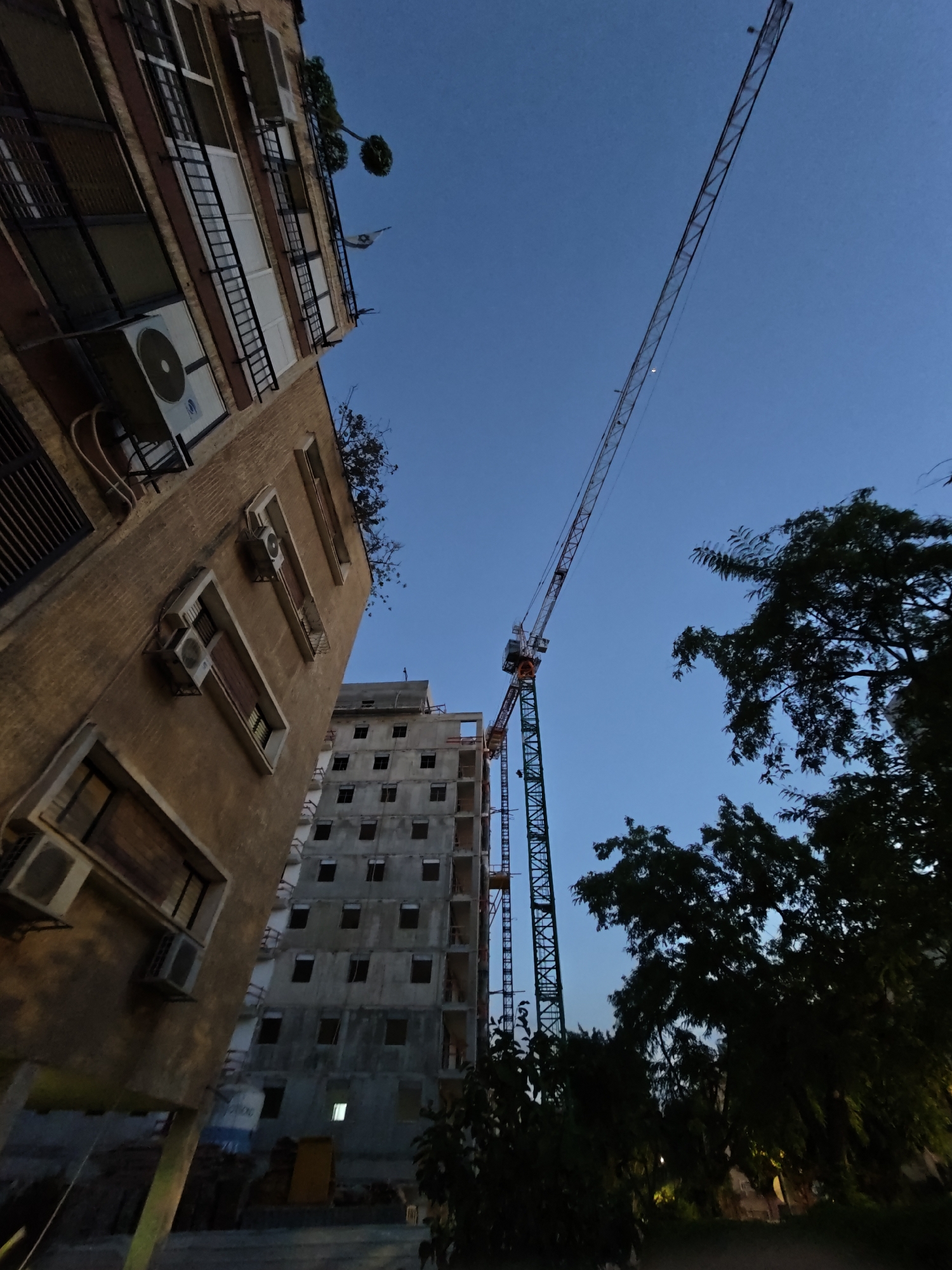 The image depicts a construction site in Petah Tikva, Israel, dominated by a tall tower crane reaching into a clear blue dusk sky. To the left, a multi-story apartment building with a brown facade and visible air conditioning units is angled towards the viewer. Rows of windows and balconies are evident on its side. In the center, a partially constructed concrete building, also multi-story, stands with its window frames visible but the glass missing, indicating an ongoing construction project. The base of the tower crane is green and lattice-like, supporting a long, extendable boom. Dark silhouettes of trees are present on the right side of the frame, with some foliage obscuring the lower part of the construction site. The scene appears to be captured from a low angle, looking upwards.
