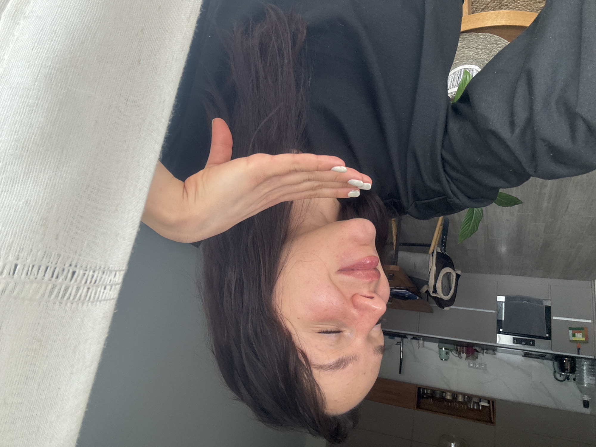 A woman with dark brown hair is positioned upside down in the image. She is wearing a black long-sleeved shirt. Her right hand is raised near her face, with white nail polish visible. The background includes a kitchen area with gray cabinets, countertops, and appliances like a microwave. A plant is visible near a wooden chair. A white curtain hangs on the left side of the frame. There is no information in the image to ascertain the location in Burgas, Bulgaria.