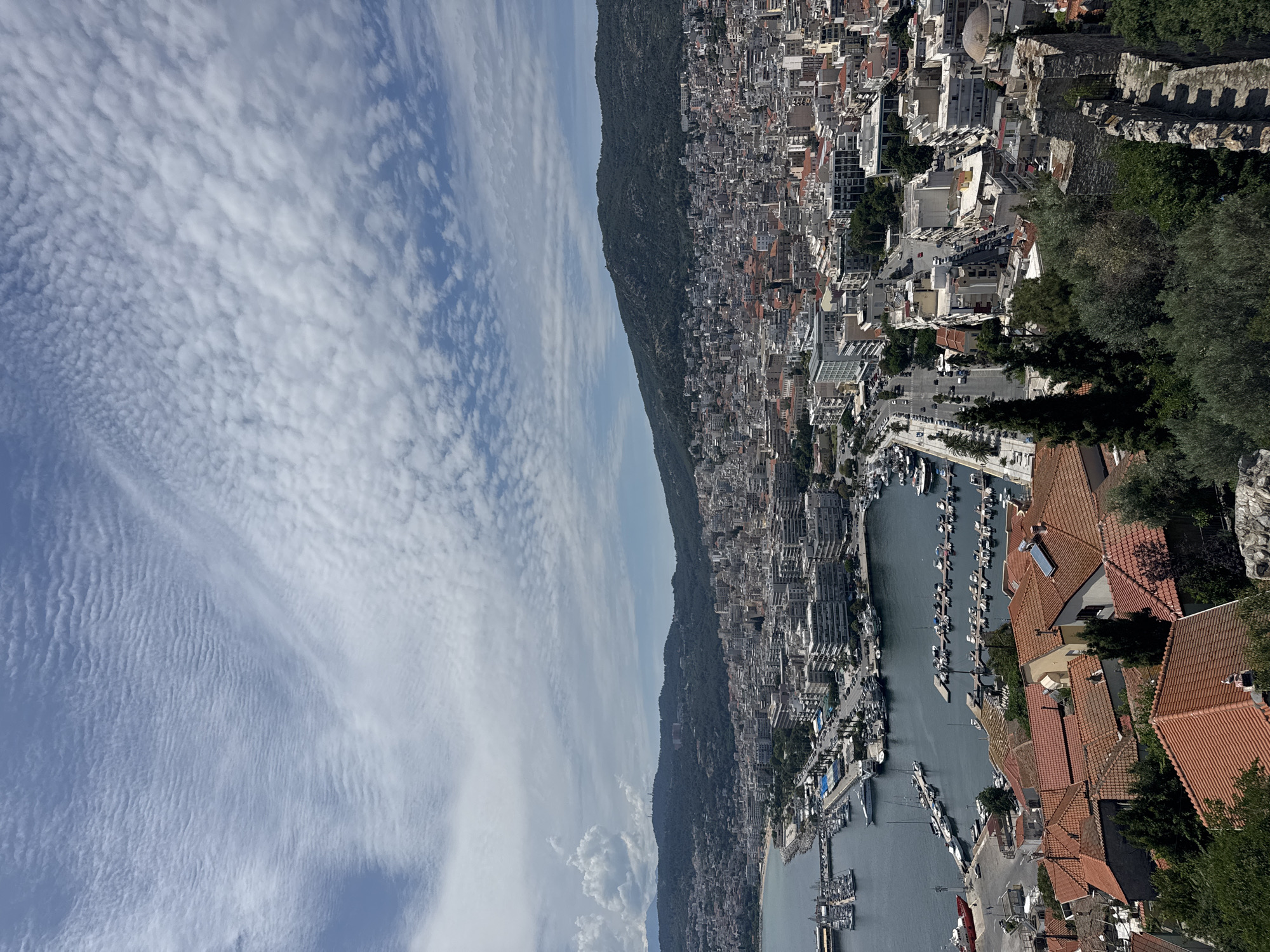 An elevated, wide-angle, full shot captures a coastal city with a port, hills, and a blue sky filled with white, rippled clouds. In the foreground, the terracotta roofs of houses are visible, interspersed with green trees and a stone wall.
The middle ground reveals a dense urban landscape of buildings with white and grey facades, many featuring balconies. A road with passing cars runs along the waterfront. To the left, a harbor is filled with numerous docked boats and yachts, and further out, larger vessels are visible.
The background is dominated by lush, green hills that encircle the city. The sky above is a bright blue, with a pattern of scattered, wavy white clouds. The overall scene depicts a sunny day in a bustling port city nestled against a natural landscape.