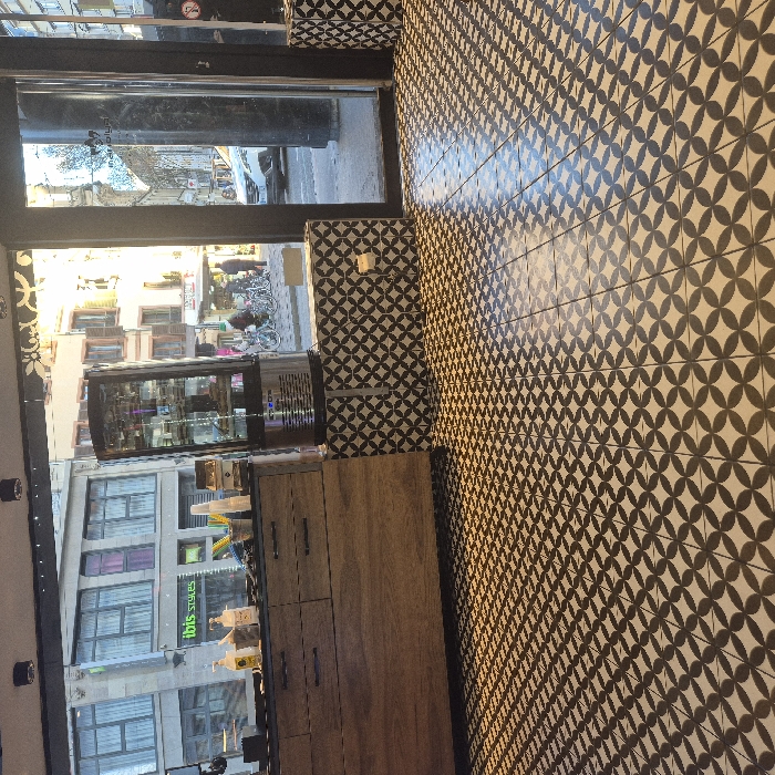 A café or small restaurant interior is shown. Black and white patterned tiles cover the floor and wall. A wooden cabinet with several drawers stands next to a glass display case. Outside, a street scene unfolds, including buildings, people, bicycles, and parked cars. An "Ibis Styles" sign is also visible through the window. The view is from inside looking out.