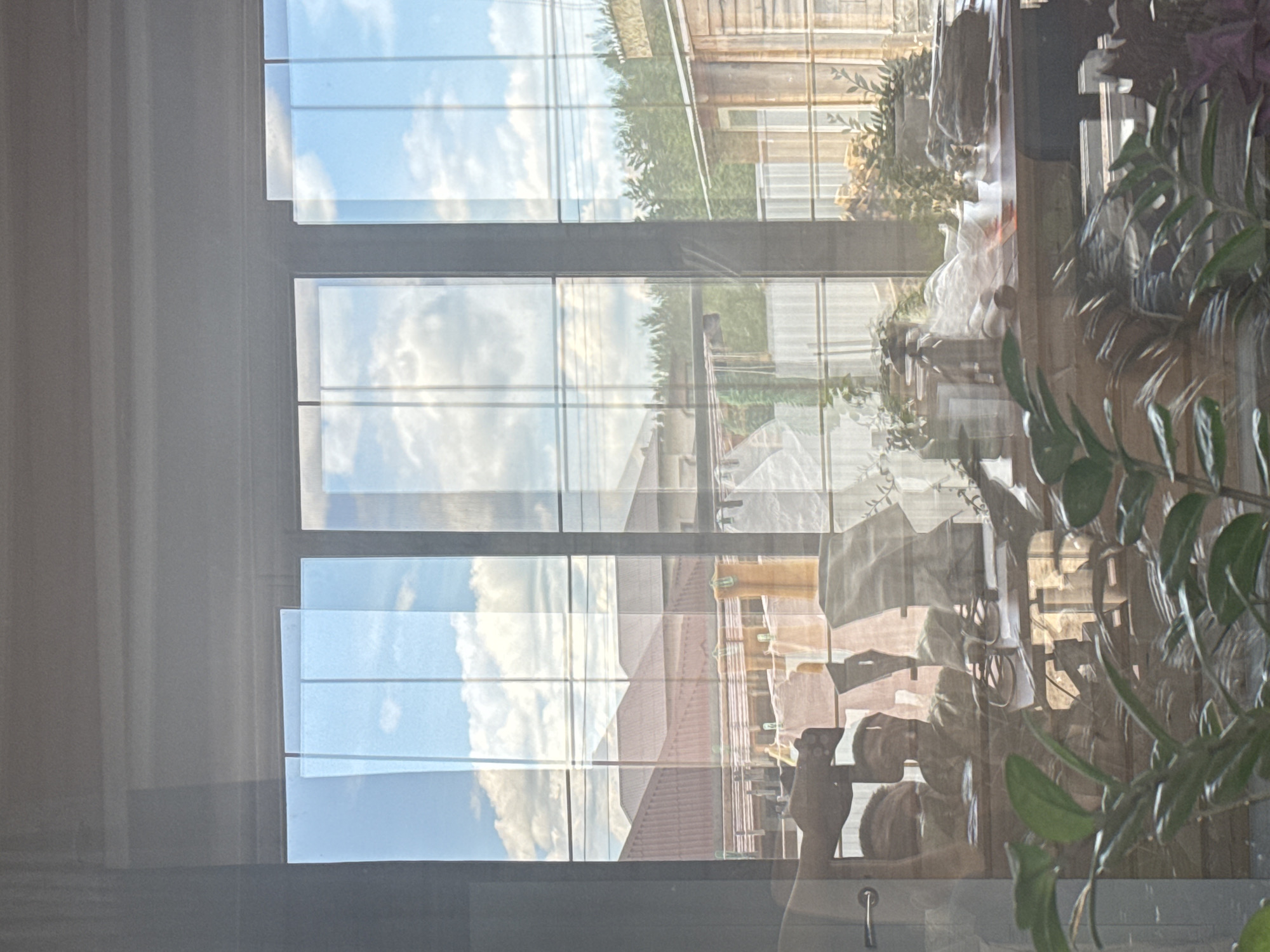 The image shows an interior view through a window with a plant in the foreground. Outside, a building with a red roof and another with wooden cladding are visible under a blue sky with clouds. Reflections of objects within the room are superimposed on the outdoor scene, creating a blurred effect. The location is Unknown, Unknown.