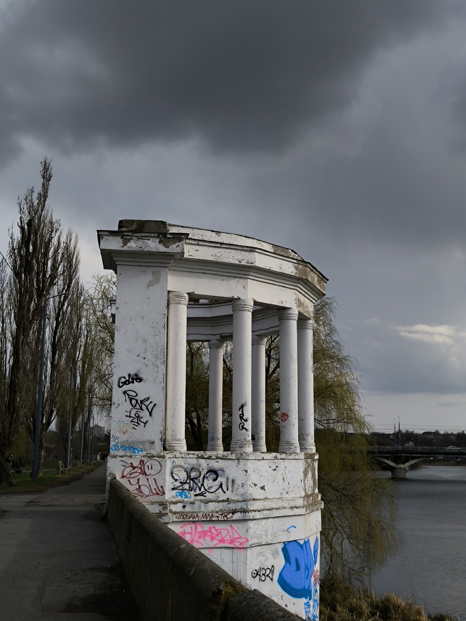 The image captures a white, derelict pavilion adorned with graffiti, located on the banks of the Southern Bug River in Vinnytsia, Ukraine. The pavilion's structure comprises pillars supporting a dilapidated roof. The walls are covered in colorful graffiti tags. A stone railing runs alongside the pavilion, separating it from a pedestrian walkway. Bare trees line the walkway, contrasting with the gray sky above. In the background, a bridge spans the river, connecting the cityscape. The overall scene conveys a sense of urban decay and neglect within a natural environment.