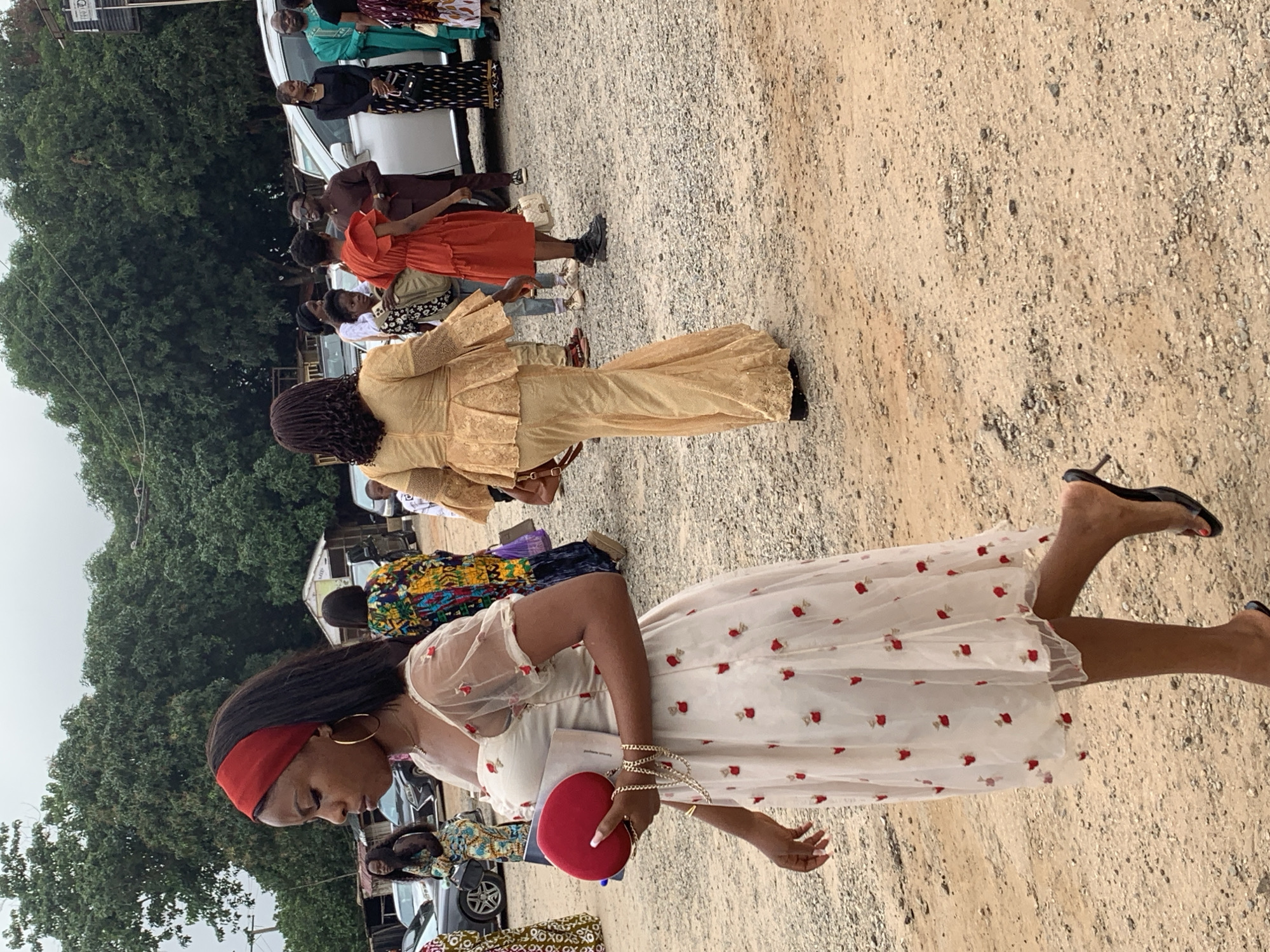 In an outdoor, gravelly area in Abuja, Nigeria, a Black woman with a red headband and gold hoop earrings walks towards the left. She wears a sheer, off-white dress with red floral embroidery and puff sleeves. She carries a red clutch in her left hand and a book in her right, along with a gold chain bracelet on her right wrist. She is wearing black, open-toed heeled sandals. In the background, several other Black individuals are visible, some wearing colorful traditional African attire. Cars are parked in the background, suggesting a gathering or event. Trees and a cloudy sky form the backdrop.