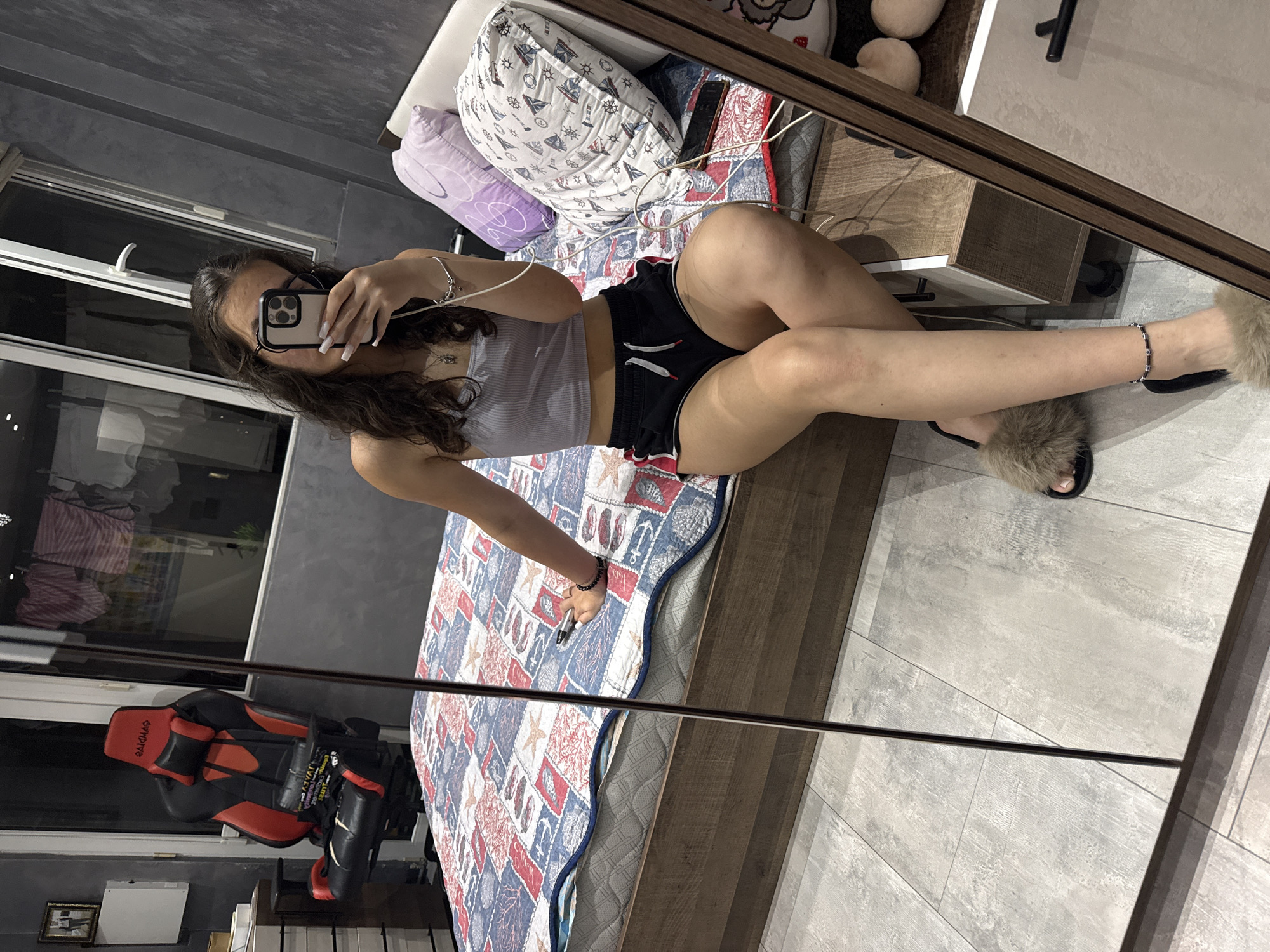 A young woman is taking a mirror selfie in a bedroom. She is sitting on the edge of a bed with a patterned quilt. She is wearing a grey tank top and black shorts. She has fluffy brown slippers on her feet and an anklet. In the background, there is a gaming chair, a dresser, a window with clothes drying on a line, and pillows on the bed. A phone is held in her right hand, and she appears to be using it to take the photo. The floor is tiled. The scene suggests a casual, domestic setting.