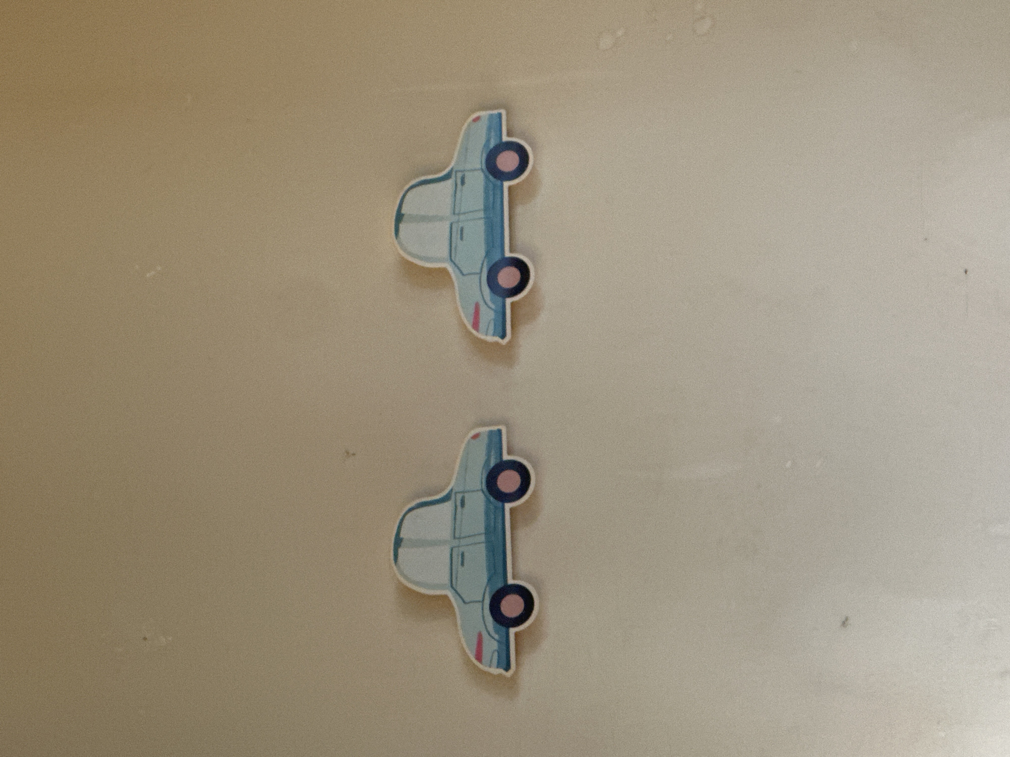 Two blue car-shaped magnets are attached to a beige or light brown wall. The magnets are vertically aligned, one above the other, with a space between them. The cars are facing to the right, with pink details on the wheels and body. The background is a plain wall with a slight texture.