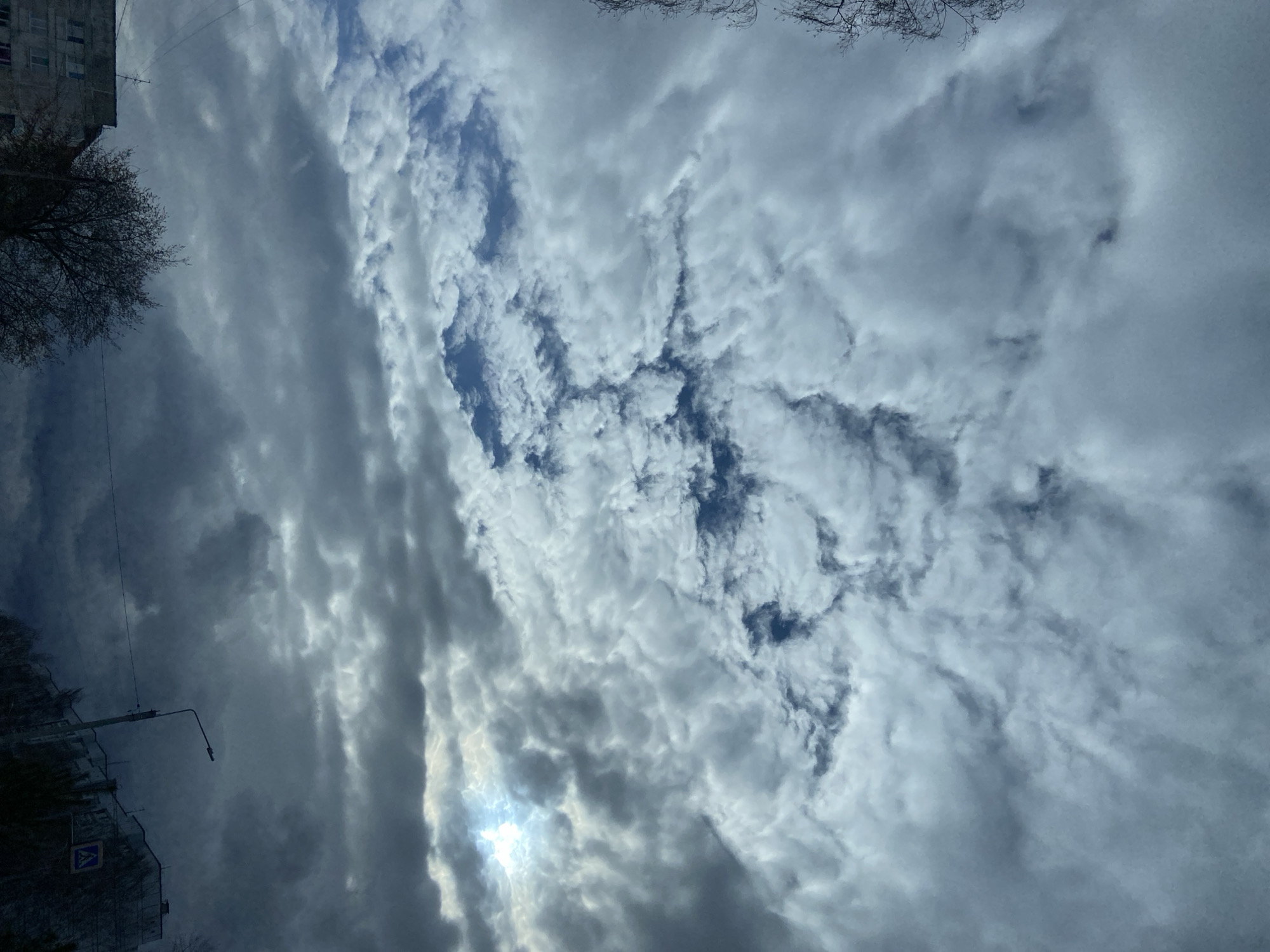 The image presents a dramatic sky filled with an intricate pattern of clouds over an urban or suburban environment, consistent with a location like Chaikovsky, Russia. The photograph is oriented vertically, with the sky dominating the central and right portions of the frame.
The clouds display a wide range of forms and shades, from bright, textured white masses with clear patches of blue sky visible, to darker, more uniform gray layers. In the lower-right section of the image, a bright, luminous spot indicates the sun partially obscured by the clouds, casting a soft, radiant light that emphasizes the depth and texture of the cloudscape. Some cloud formations appear dense and layered, while others show more defined, rippled, or altocumulus-like structures, particularly towards the center and upper right.
On the left side of the frame, elements of the ground-level environment are visible. In the upper left corner, a section of a multi-story building, likely residential, with visible windows, can be seen. Below the building, the silhouetted, bare branches of a tree extend into the sky. Further down on the left, a street light or utility pole with an overhead arm and wires is present. Attached to this pole is a blue pedestrian crossing sign, depicting a white triangle with a black walking figure.
The scene depicts daytime conditions, characterized by the interplay of sunlight and varying cloud cover, suggesting a partly cloudy or overcast day with moments of brightness. No specific human activity is captured, focusing instead on the atmospheric conditions and the static urban details.
