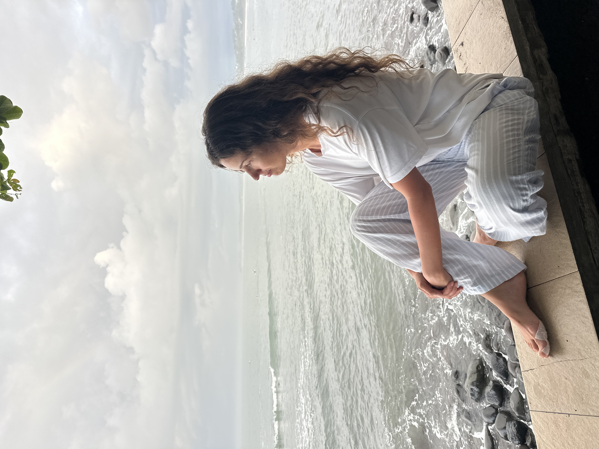 The image captures a woman in Pekutatan, Indonesia, kneeling on a tiled surface bordering a rocky shoreline where ocean waves break. She is dressed in a white t-shirt and light-striped trousers. Her long, curly brown hair cascades down her back as she leans forward, gazing downward with her hands clasped in front of her. The background features a grey, overcast sky meeting the horizon of a pale, rippling ocean. A small patch of green foliage is visible in the upper left corner.