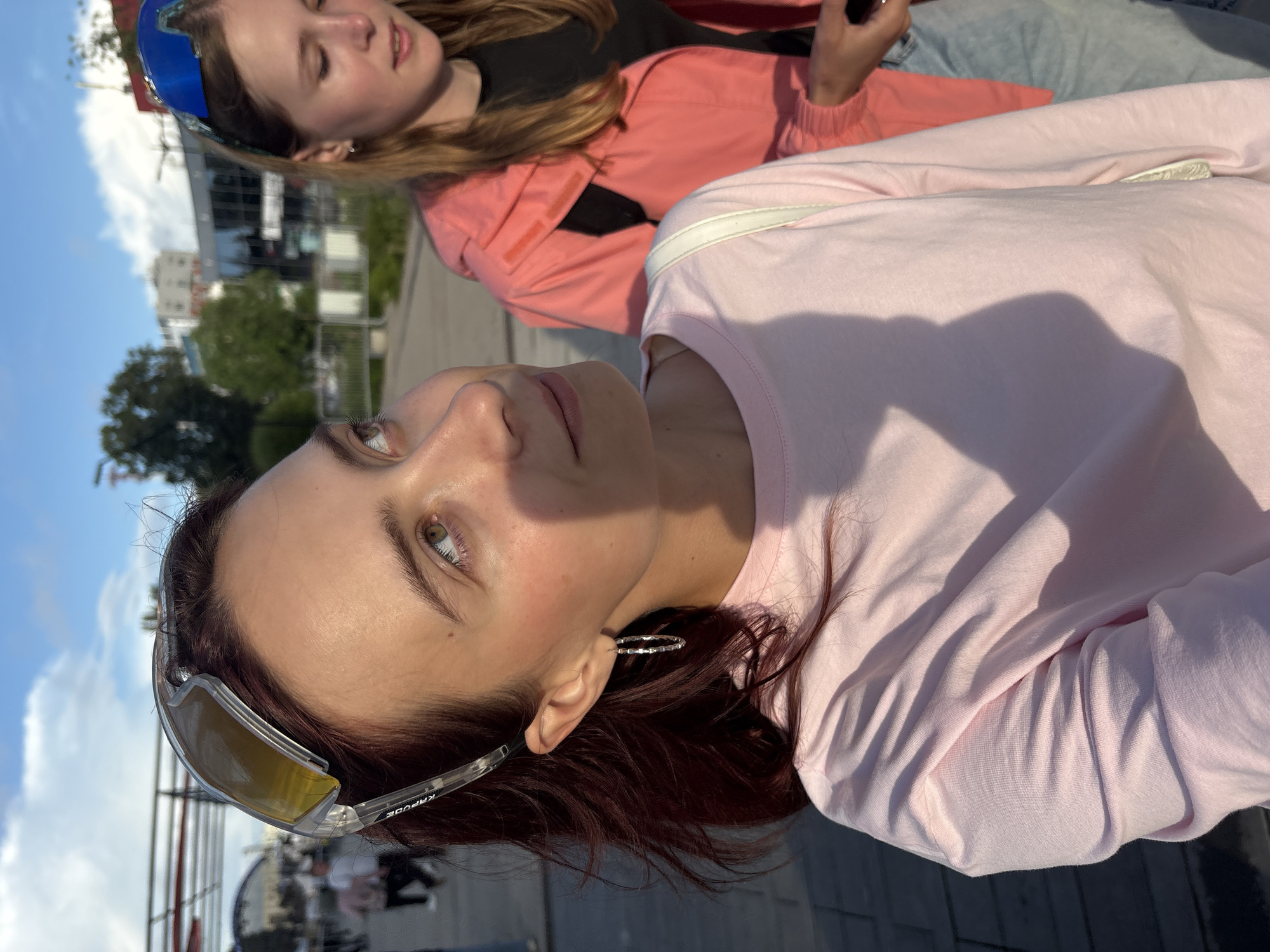 The image shows two young women outdoors on a sunny day, likely in a public area of Izhevsk, Russia, based on the context provided. The woman in the foreground, with reddish-brown hair, is wearing a pink long-sleeved shirt and large, clear-lensed sports sunglasses pushed up onto her head. She has a silver hoop earring in her visible ear and is looking up and to her right, with a slight smile. A white strap from a bag is visible over her shoulder.
Behind her and slightly to the right, another young woman with blonde hair is wearing a bright pink hooded jacket over a black top and light blue jeans. She has blue sunglasses perched on her head and is looking downwards, holding a black smartphone in her right hand.
The background suggests an urban environment with buildings, trees, and people in the distance. The lighting indicates it is daytime, with shadows cast by the people and overhead structures. The ground appears to be paved. The overall scene depicts a casual outing between friends.