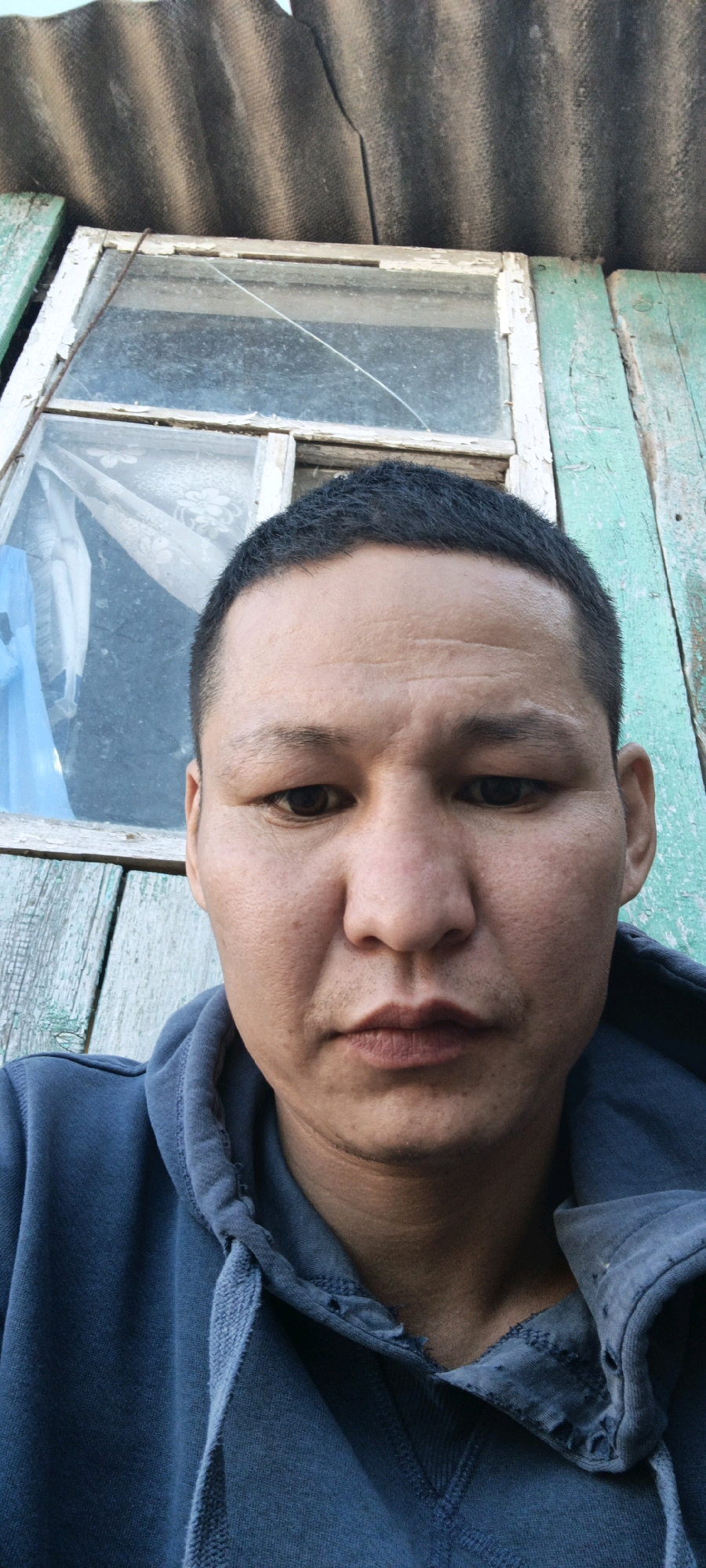 A close-up, front-facing selfie shows a man's face, from his hairline to his upper chest. He is wearing a dark blue hooded sweatshirt. The man has short, dark hair, dark eyes, and a neutral expression. His skin appears tanned, with some visible pores and minor blemishes. He has light stubble on his upper lip and chin.
Behind the man, a rustic window frame with peeling white paint is visible. The window glass is dirty and has a crack running diagonally across it. Part of a blue tarp or plastic sheeting is visible inside the window. The window is set into a wall made of weathered, pale blue wooden planks. Above the window and the wall, a section of a corrugated, gray roof made of asbestos cement sheets is visible. The scene suggests an outdoor setting, possibly a rural or dilapidated structure. The overall impression is one of a candid, unposed moment.