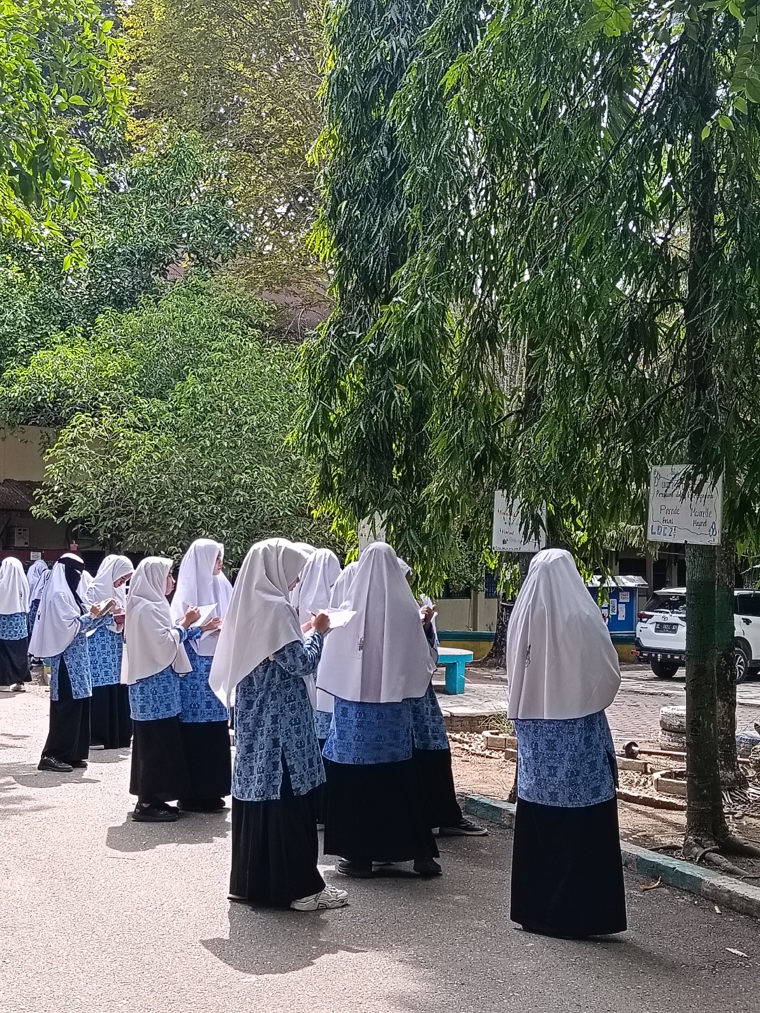 In Lhokseumawe, Indonesia, a group of students, likely female given their attire, stand in a line on an asphalt surface. They are all wearing white hijabs and blue patterned uniforms with black skirts. Most of the students are holding notebooks or papers and appear to be writing or looking at something in their hands. They are standing outdoors, with lush green trees overhead and to the sides, suggesting a park or school grounds. In the background, there are buildings, a light blue bench, and vehicles parked on a dirt area. Signs are visible on one of the trees, with some text legible, indicating it might be a public area or educational institution. The lighting suggests it is daytime, with shadows cast on the ground.