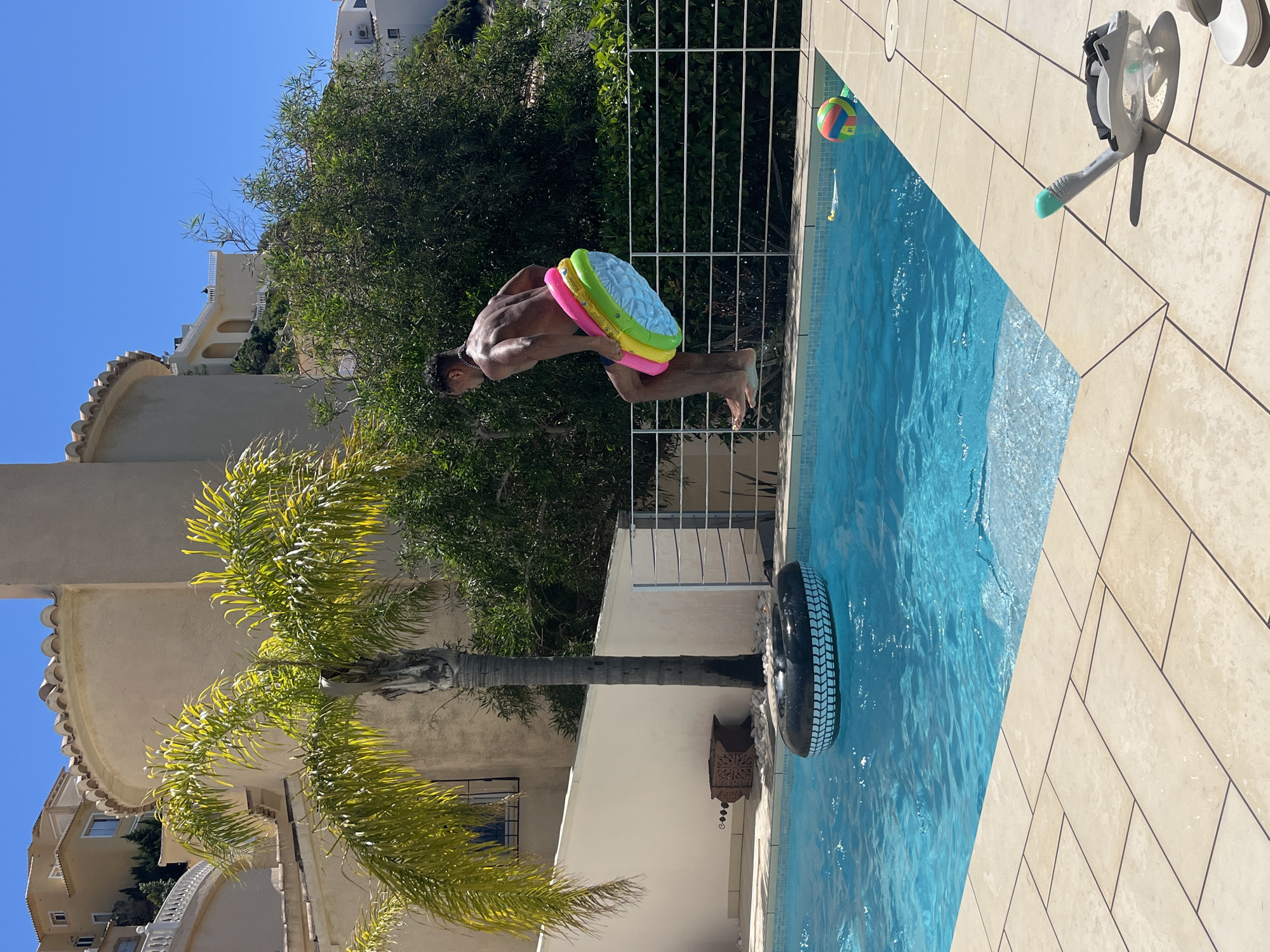 The image captures a man diving into a swimming pool in what appears to be a residential area of el Poble Nou de Benitatxell / Benitachell, Spain. The man, who is shirtless and wearing swim trunks, is in mid-air, with a colorful inflatable ring around his waist. He is positioned to enter the pool headfirst.
The pool is rectangular and filled with blue water. A black inflatable tire is floating in the water. A snorkel and diving mask are positioned on the deck of the pool. On the pool deck, the tiles are arranged in a herringbone pattern.
Behind the man, there is a white railing and dense green foliage. In the background, there are buildings, likely residential homes. The sky is clear and blue. A tall palm tree is visible to the left of the frame. The image appears to have been taken during daylight hours.