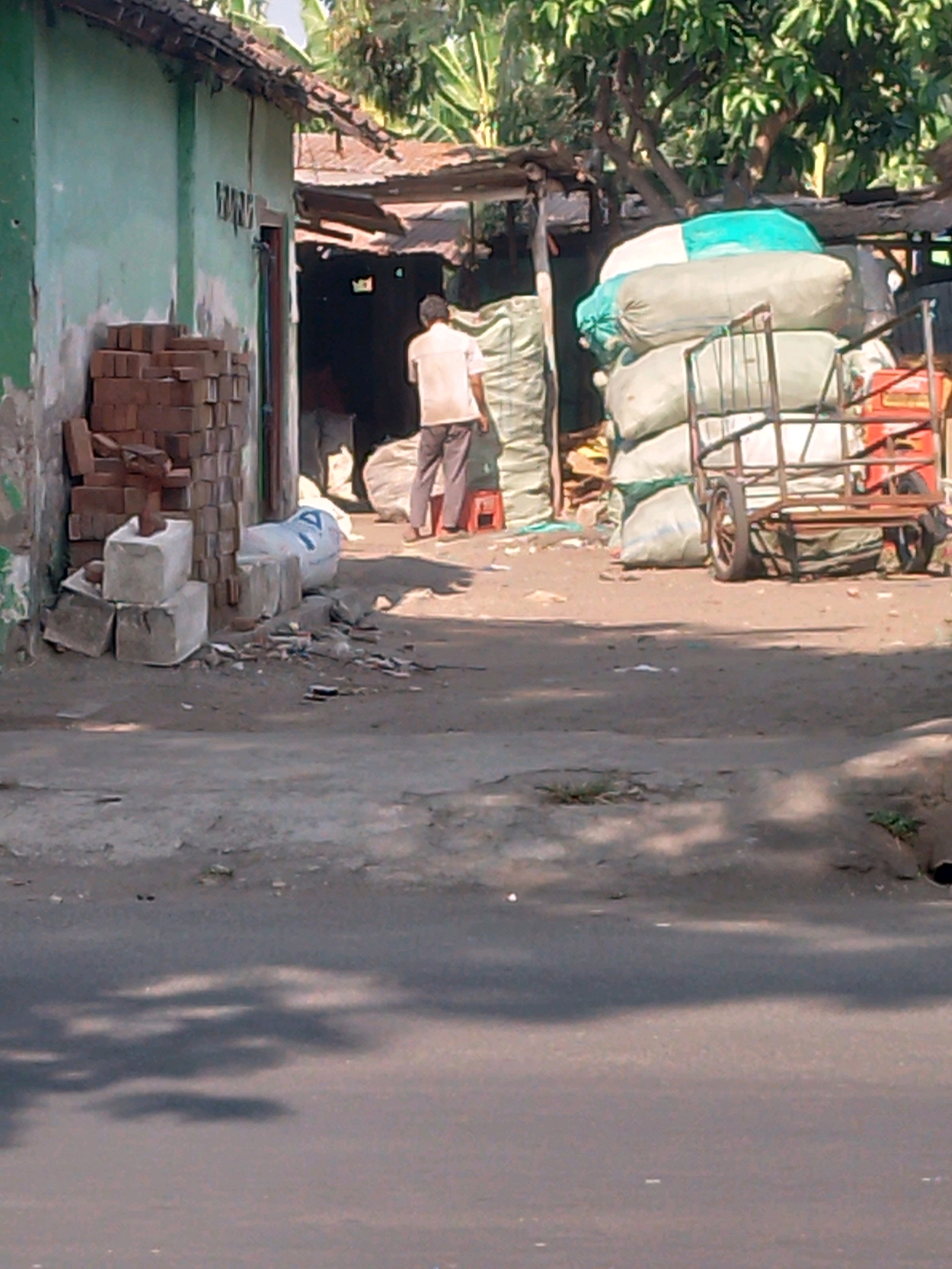 This is a mid-distance, eye-level photograph of a street scene in Kertosono, Indonesia. The scene depicts an outdoor area adjacent to a weathered green building with a corrugated metal roof. In the foreground, a paved road is visible, with shadows cast by trees on the pavement.
To the left of the building, a stack of bricks and concrete blocks are piled up. A person, dressed in a light-colored shirt and dark pants, is standing in the middle ground, facing away from the camera, near an open doorway of the building. In front of this person are several large sacks, some white and some light green, possibly containing goods or materials. To the right, a metal cart, also laden with sacks, is positioned on the dirt ground. The cart has a rusty frame and two wheels. The background consists of foliage, indicating the presence of trees. The overall impression is of a bustling but somewhat dilapidated commercial or storage area in an urban or semi-urban setting.