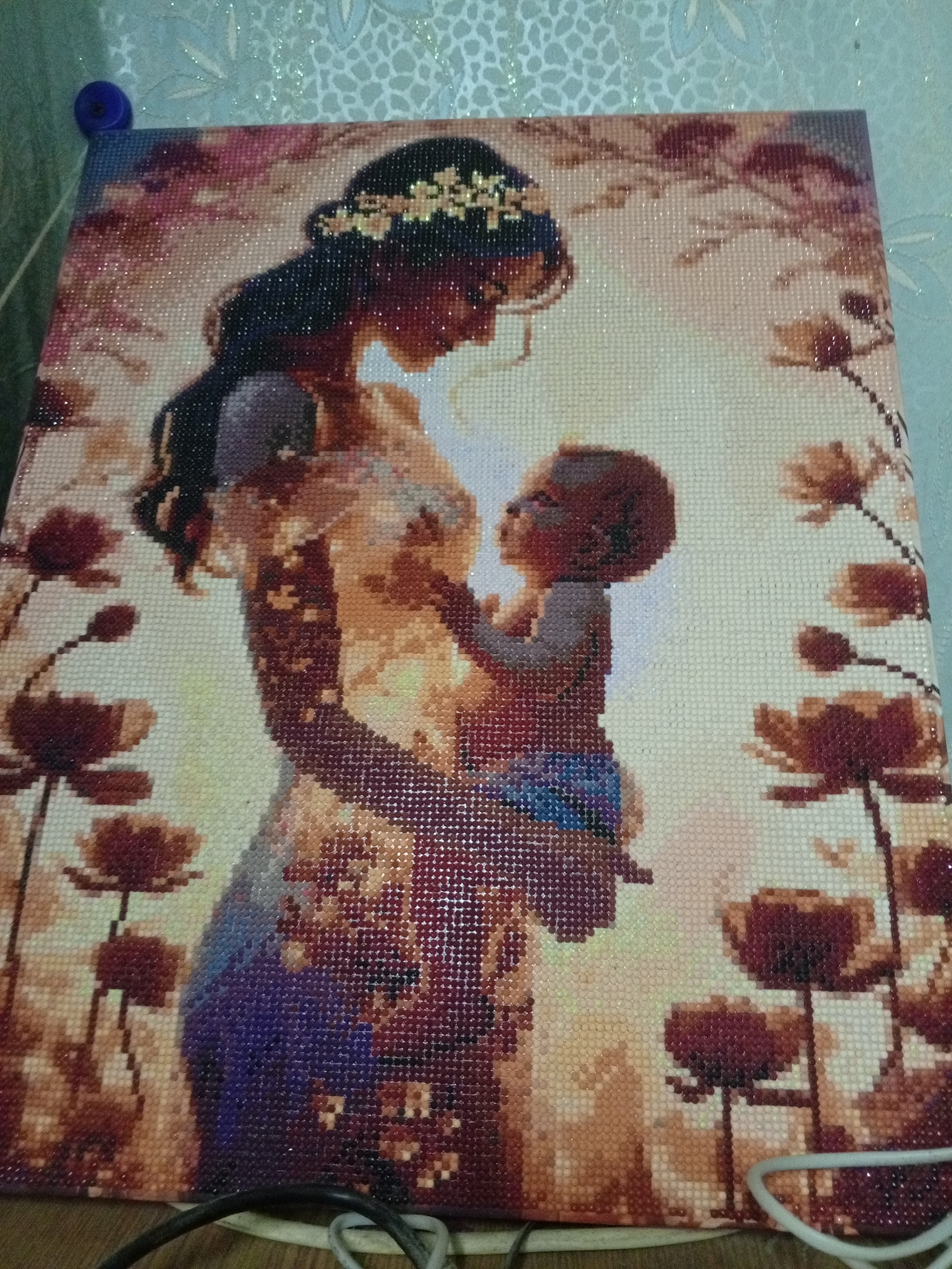 The image is a close-up photograph of a completed diamond painting depicting a mother holding a baby. The mother has long dark hair adorned with a floral headpiece, and she is looking down at the baby. The baby is fair-skinned. They are surrounded by a field of flowers. The diamond painting is made of small, faceted resin pieces. The background is a slightly blurred light blue with a subtle floral pattern. The picture was taken indoors. The exact location in the city of Puchezh, Russia, is not discernible from the image.