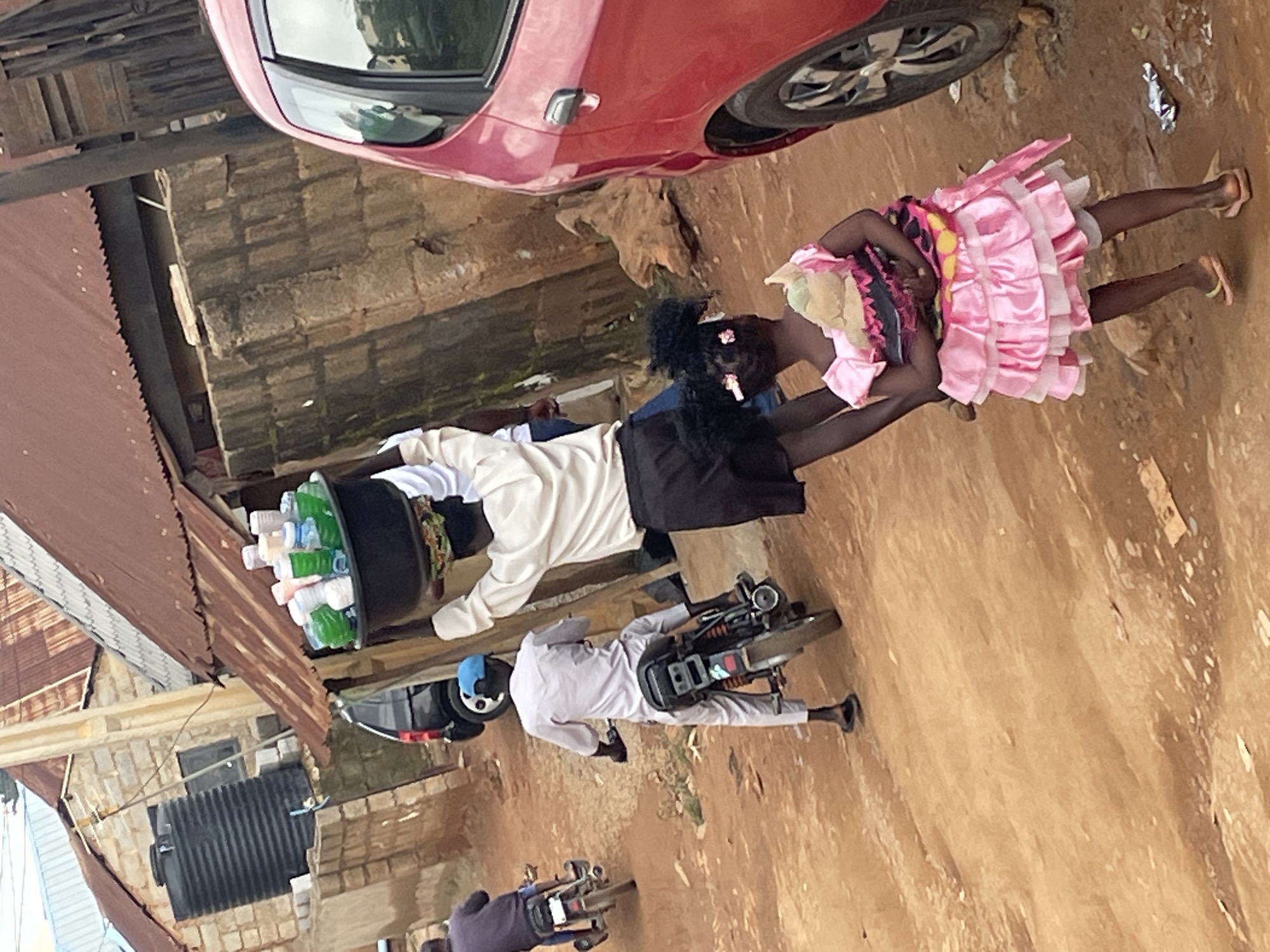 In Kurunduma, Nigeria, a person is walking on a dirt road, balancing a large black bowl filled with plastic containers and bottles on their head. The bowl contains numerous items, including white and green plastic cups, and several bottles, some appearing to be water bottles. The person is wearing a white long-sleeved shirt and dark shorts, with a patterned cloth wrapped around their waist. Behind this person, a young girl in a pink dress with ruffled layers and a doll is walking. Two individuals on motorcycles are visible in the background; one is wearing a light-colored outfit and a blue cap, while the other is wearing a dark shirt. A red car is parked on the right side of the image. The environment is characterized by unpaved ground, buildings constructed from concrete blocks with corrugated metal roofs, and a large black water tank attached to one of the structures.