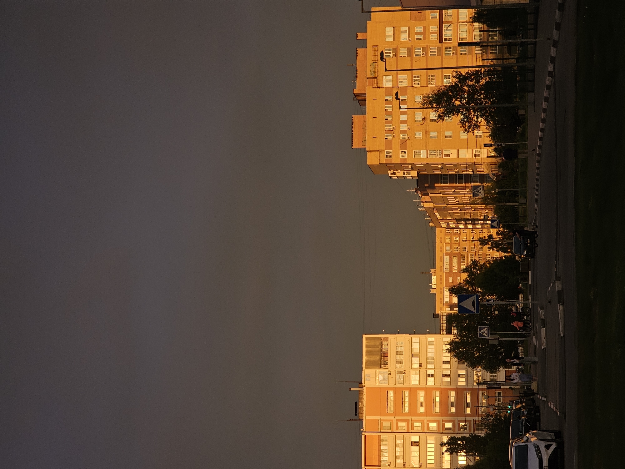 The image depicts an urban scene in Nizhny Novgorod, Russia, during sunset. The sky is a gradient of dark gray to a warm, golden hue, illuminated by the setting sun. In the background, a row of apartment buildings stands tall, their facades reflecting the warm light. The buildings are a mix of orange and yellow tones, with numerous windows visible, some of which appear lit from within. Trees are scattered throughout the scene, adding a touch of greenery against the urban architecture.
In the foreground, a street is visible with several cars parked and driving. A white car is parked on the left side of the frame. A few pedestrians are seen walking or standing on the sidewalk and crossing the street. Road markings and traffic signs are present, indicating a street intersection. The overall atmosphere is serene, with the warm light of sunset casting long shadows and highlighting the architectural details of the buildings.