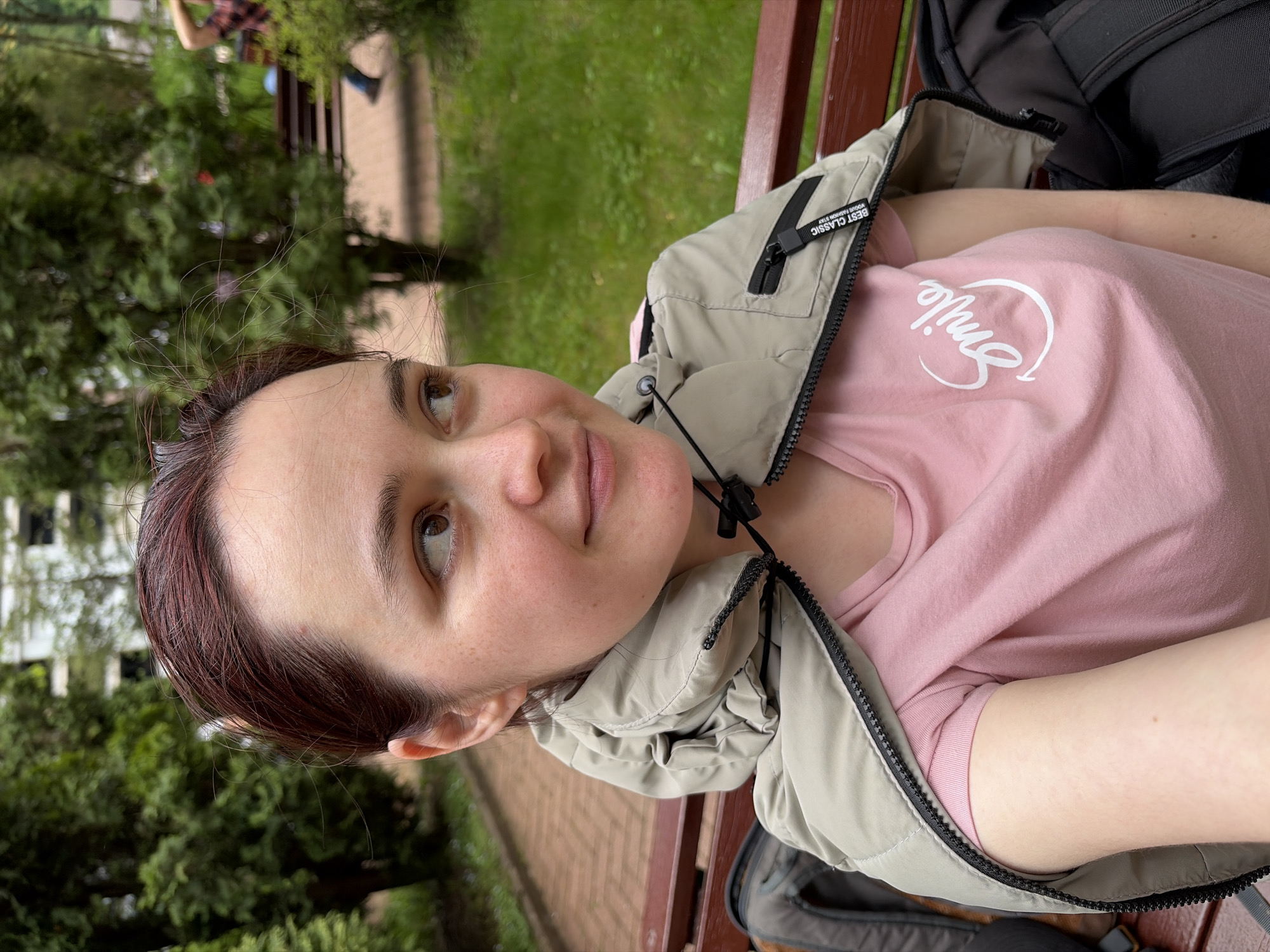 A woman is sitting on a bench. She wears a pink shirt with a "Smile" graphic and a light-colored vest. A black backpack is next to her on the bench. The setting appears to be a park or green space with trees and grass visible. In the background, there is another person sitting on a bench, and a brick walkway. Given the information about location, it is likely the photo was taken in Estosadok, Russia.
