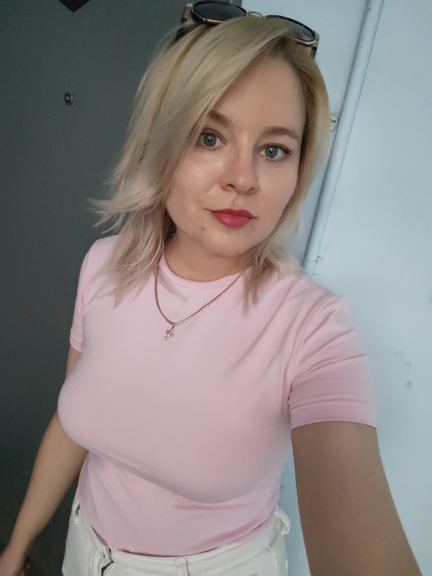 The media file is a selfie taken by a woman with blonde hair, blue eyes, and red lipstick. She is wearing a light pink t-shirt and white shorts. She has a silver necklace with a cross pendant. Sunglasses are perched on top of her head. The background appears to be indoors, with a gray wall on the left and a light-colored wall or door on the right. A portion of a doorway or doorframe is visible on the far left. The lighting suggests it might be daytime, possibly indoors near a window or with artificial lighting. The woman's expression is neutral. There is no visible text in the image. The location context is Izhevsk, Russia.