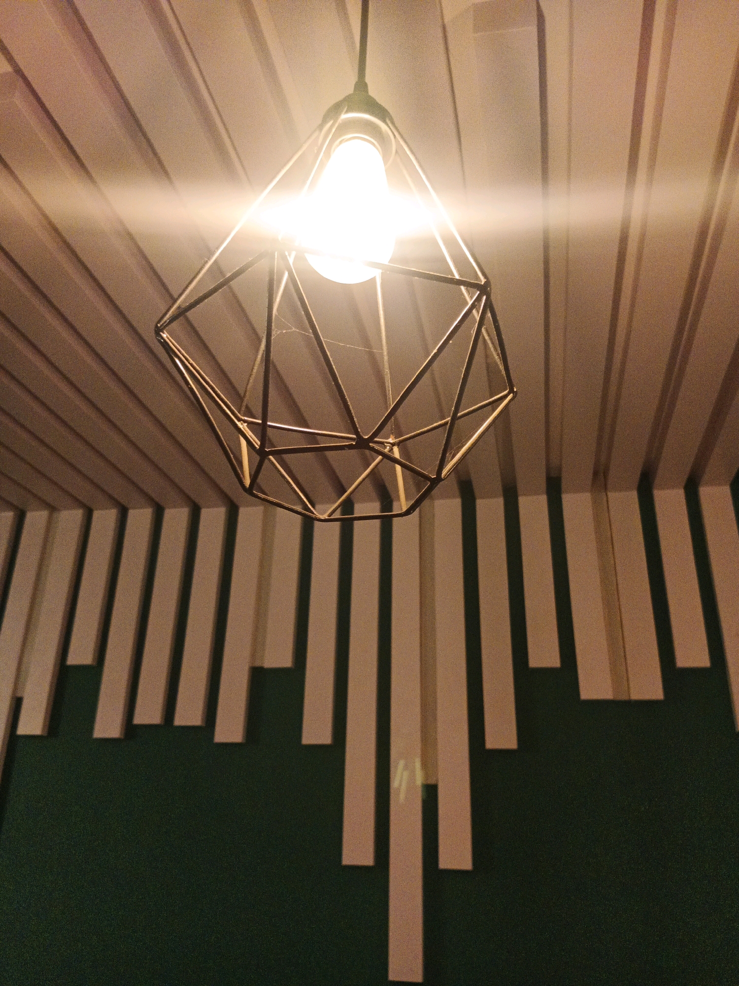 An interior shot captures a modern geometric pendant light fixture hanging from a ceiling. The ceiling is adorned with parallel wooden slats, creating a ribbed texture. Below the ceiling, the wall features a dark green textured surface. Mounted on this wall are vertical light-colored rectangular panels of varying heights, arranged in a decorative pattern, casting shadows on the green wall. A bright light bulb is illuminated within the geometric metal cage of the pendant light, casting a warm glow and creating lens flare effects. The image composition focuses on the interplay of light, shadow, and architectural elements. No people are present in the scene. The location is not specified in the image itself, nor is there any indication of Bandung City, Indonesia.
