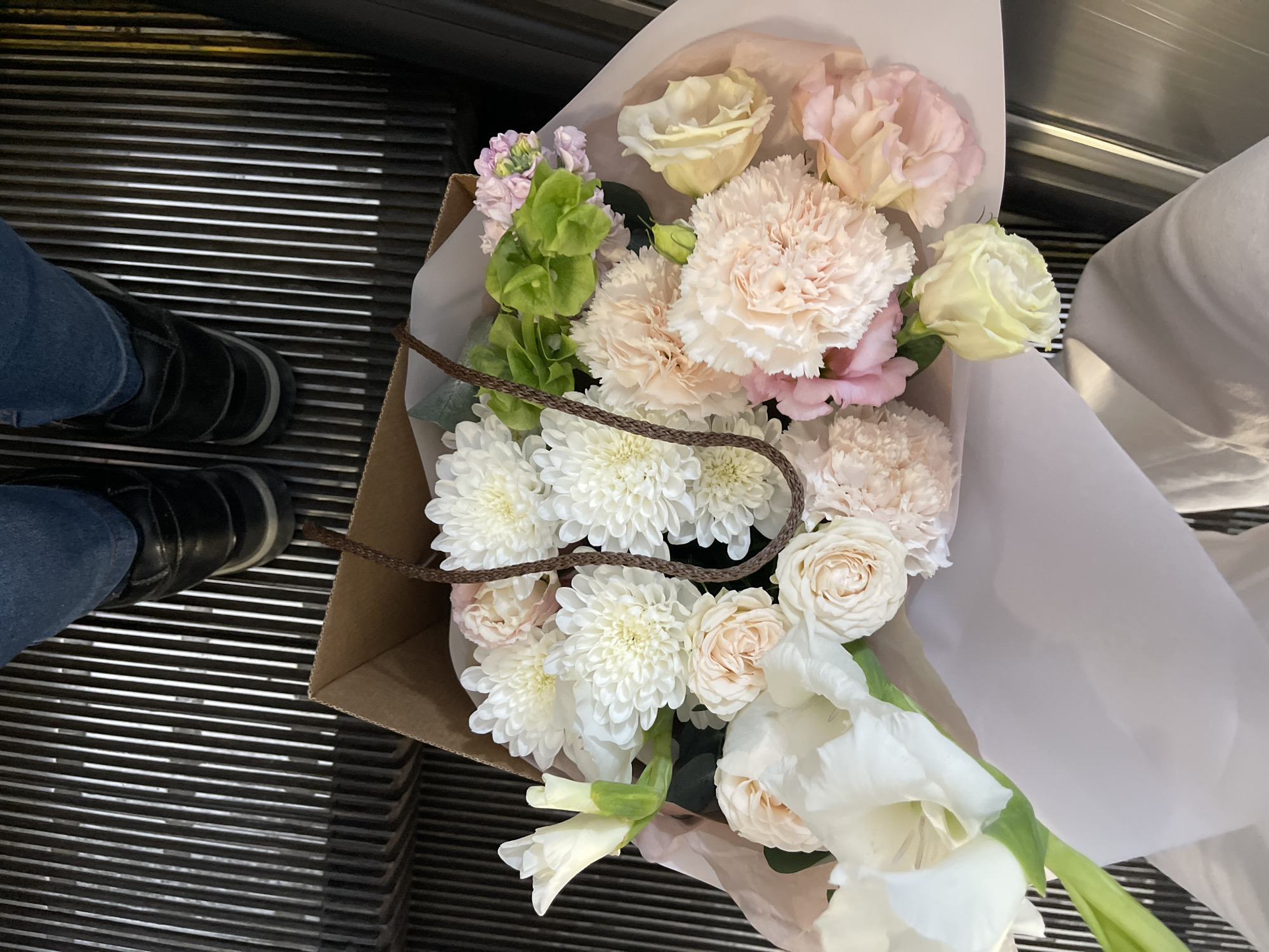 The image displays a top-down perspective of a person's lower body and a bouquet of flowers on an escalator. The individual wears blue denim jeans and black shoes with dark soles. They are holding a brown paper bag containing a large, mixed floral arrangement. The bouquet primarily consists of numerous white, dense-petaled chrysanthemums, several pale pink carnations, and light cream or blush-colored garden roses. A prominent white gladiolus flower with a green stem is visible on the left side, along with some light green foliage and small purple/pink floral accents. The flowers are wrapped in light-colored paper within the brown bag, which features a braided dark brown handle. The escalator's steps are constructed from metal treads with distinctive parallel silver-colored grooves. No specific visual cues or markers are present within the image to identify the location as Moscow, Russia.